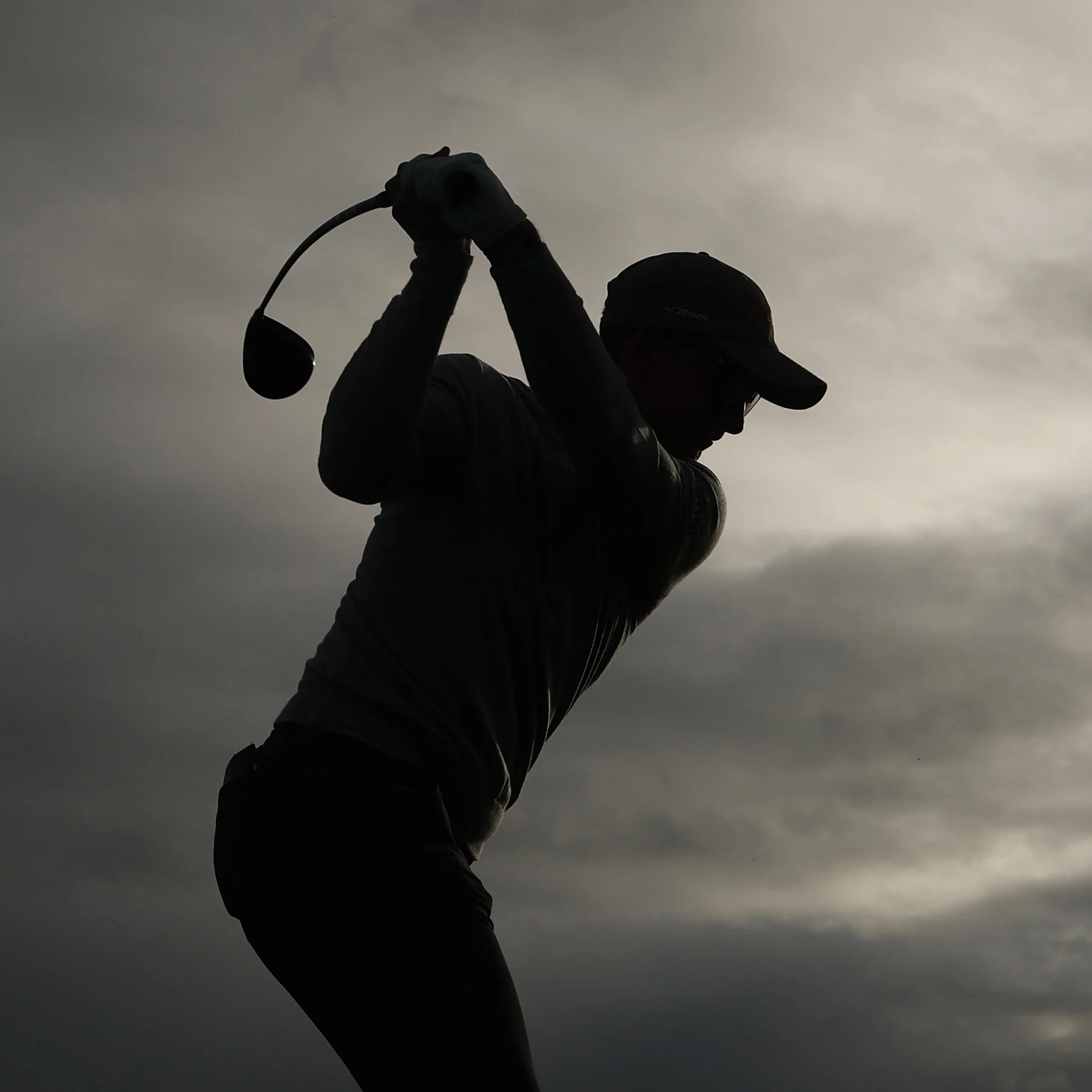 A silhouette of a male golfer mid-swing, set against a cloudy sky.