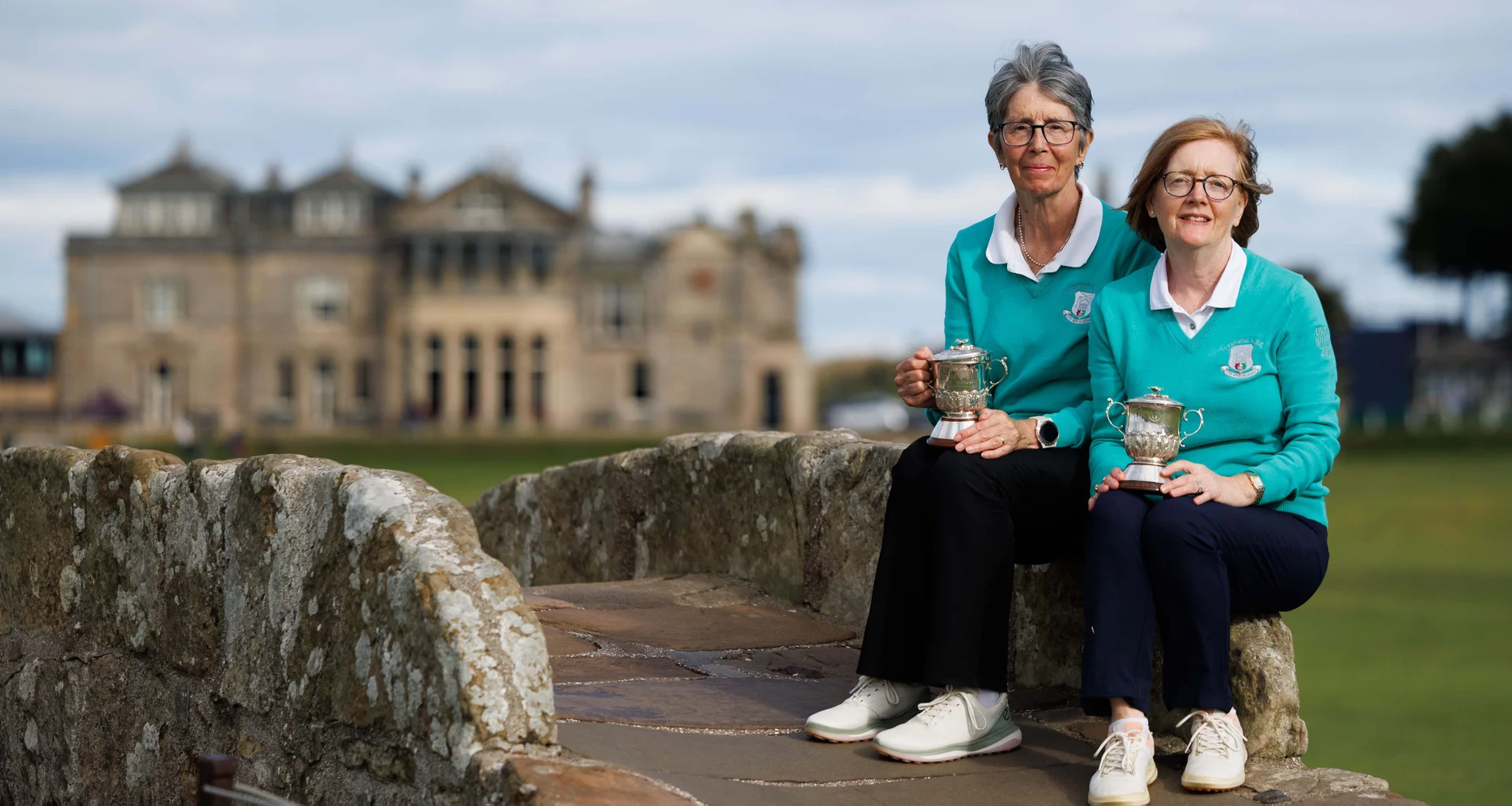 Lynn Coote and Lorraine Abbott sit on the Swilcan bridge with their trophies.