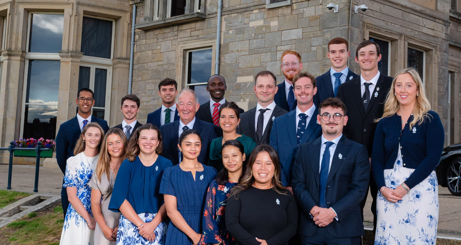 A group of people stand together looking towards the camera smiling with a building in the background.