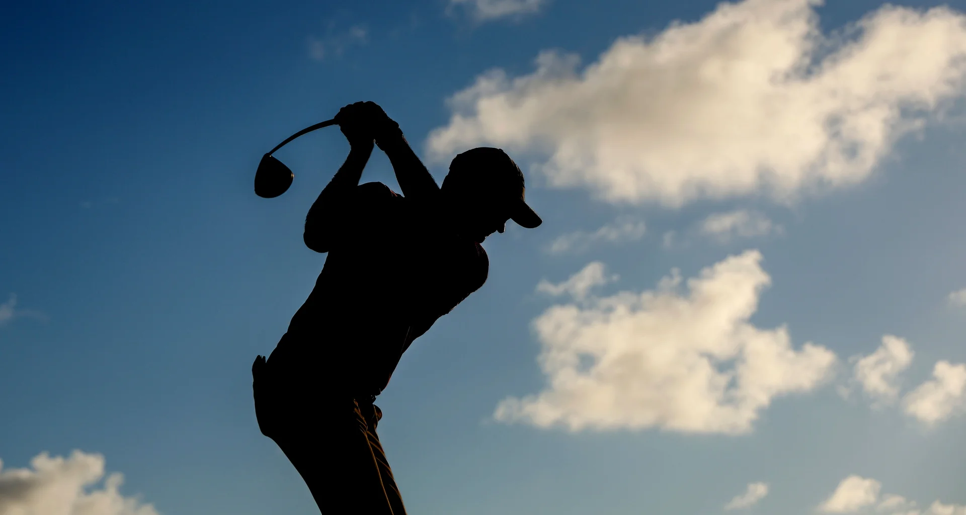 A silhouette of a male golfer teeing off with a driver set against blue sky.