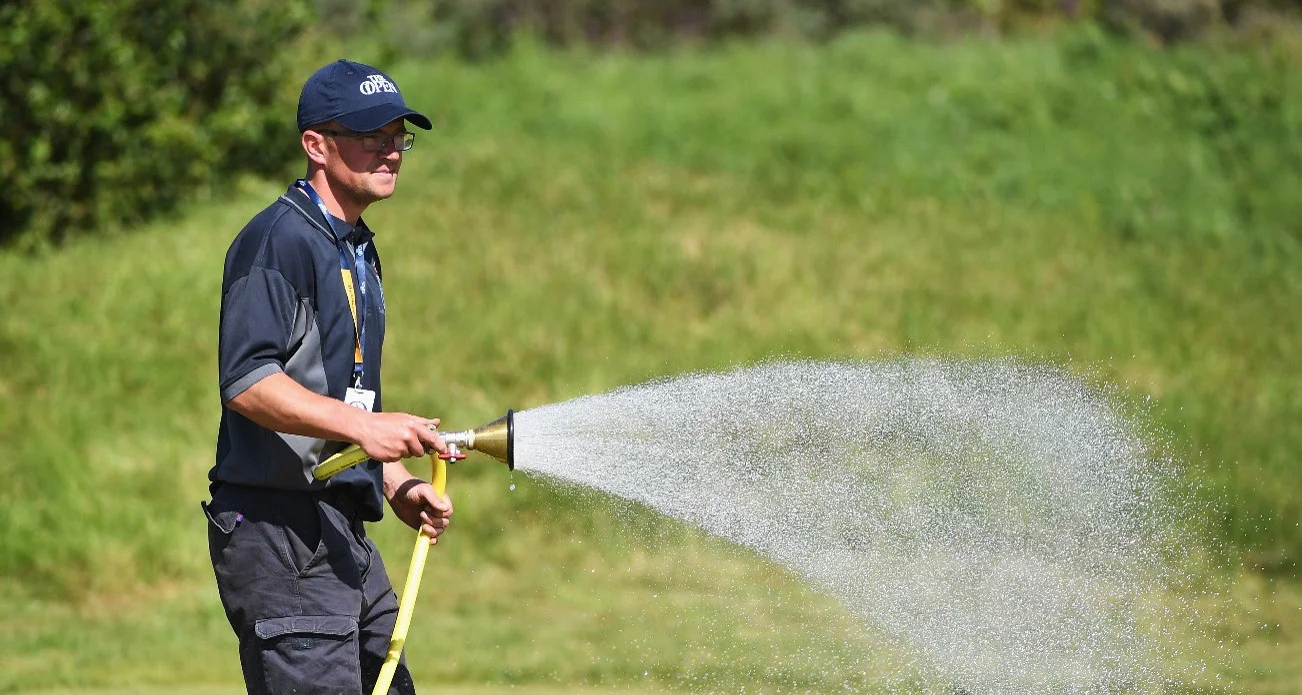 A man wearing an Open baseball cap waters a green on a golf course.