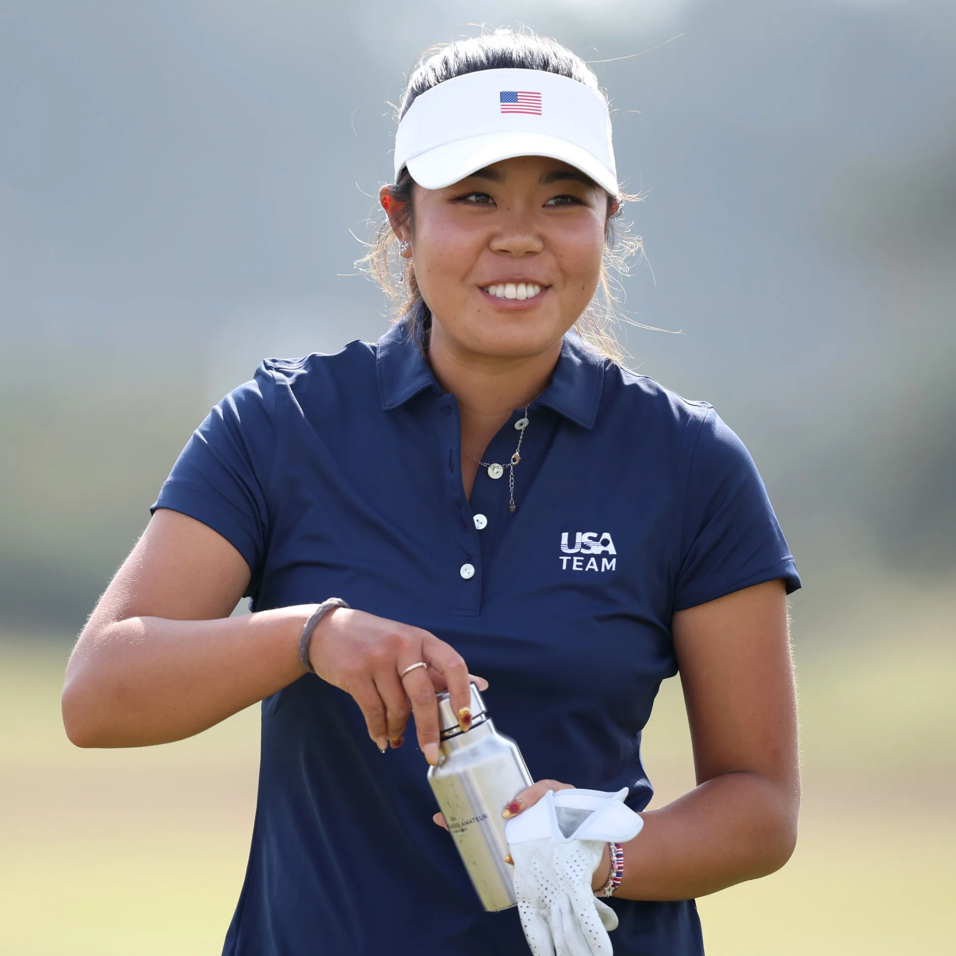 Golfer Jasmine Koo smiles brightly while handling a water bottle and wearing a white visor with a US flag, a navy blue USA Team polo, and a white golf glove.