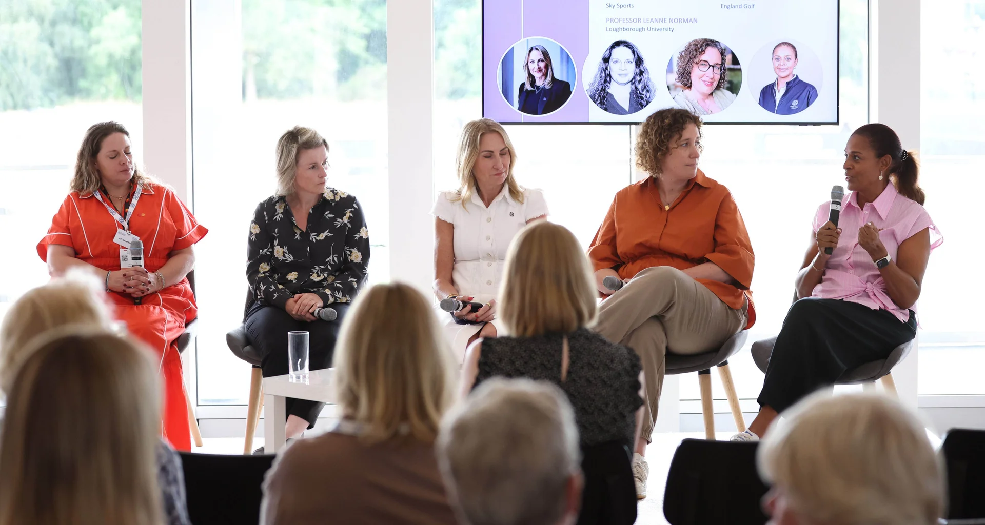 A five-woman panel sit facing the audience during the Women In Sport Summit. The person on the right holds the microphone while the other four women and audience look on.