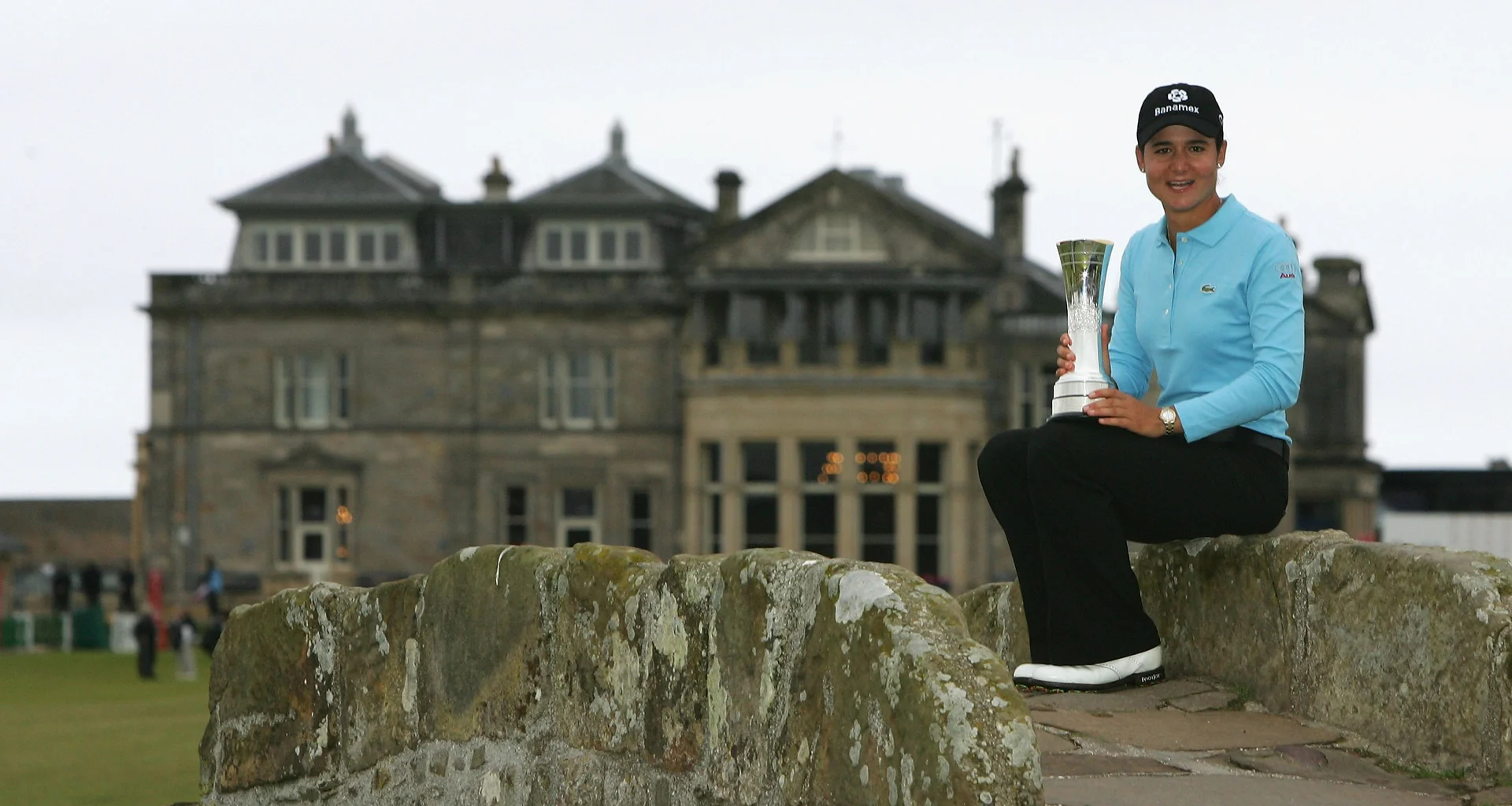 Lorena Ochoa sits on the Swilcan Bridge holding the AIG Women's Open trophy with The Royal and Ancient Golf Club of St Andrews in the background.