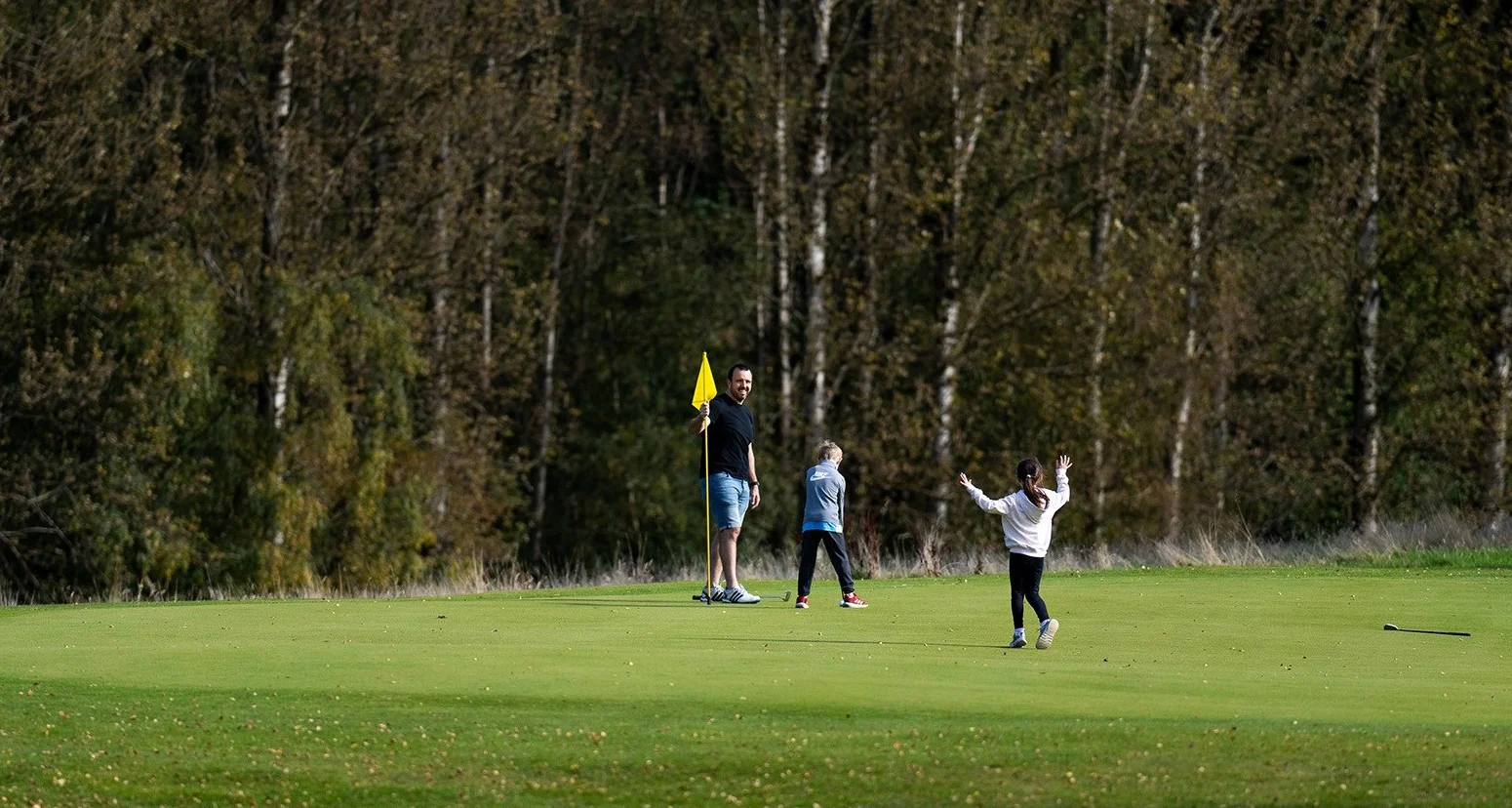 A man and a young boy and girl stand happily on a golf green. The man hold the pin flag upright while the boy plays a shot.