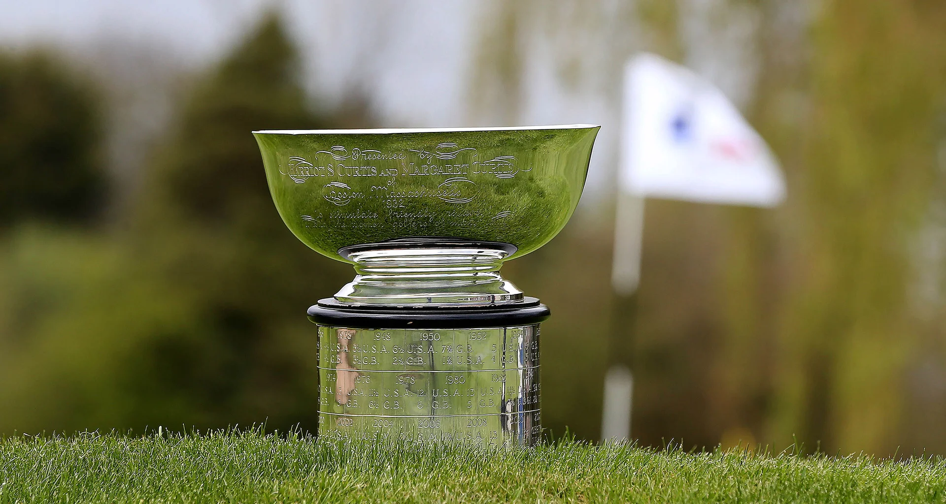 The Curtis Cup trophy sits on grass beside a golf green. A white pin flag is visible but out of focus in the background.