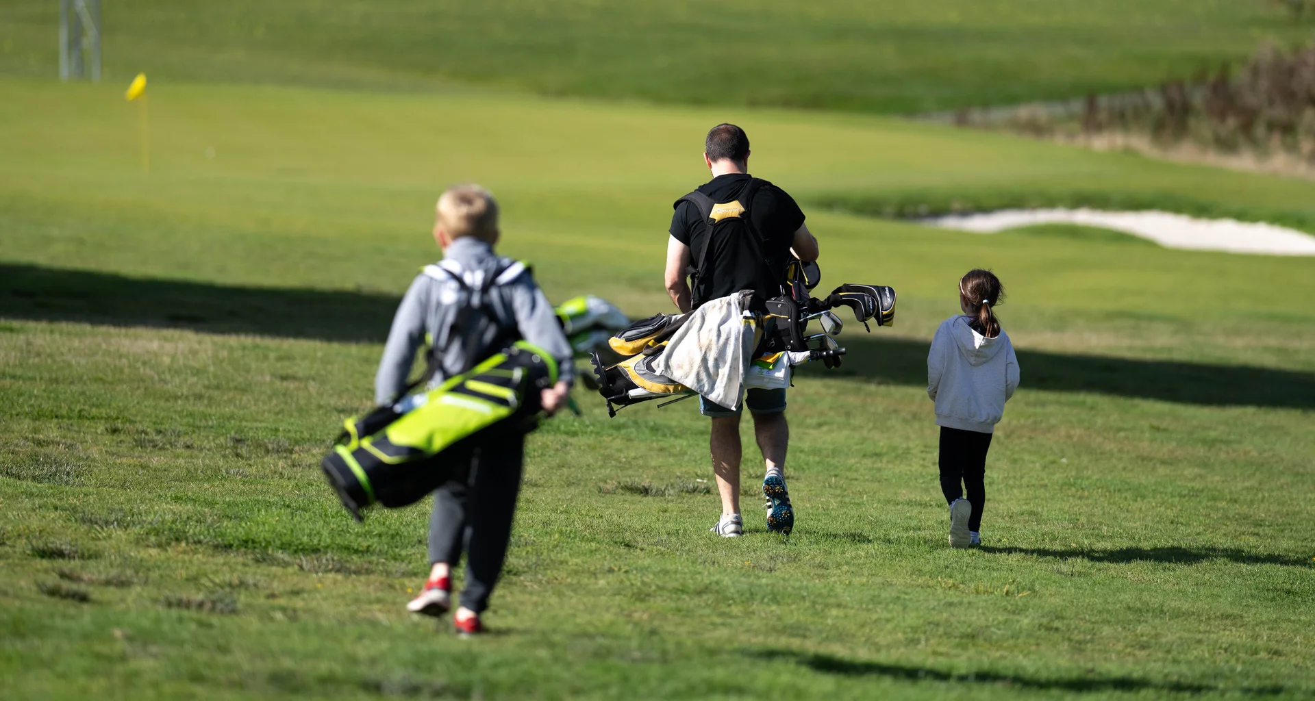 A man and two children walk on a fairway carrying golf clubs.