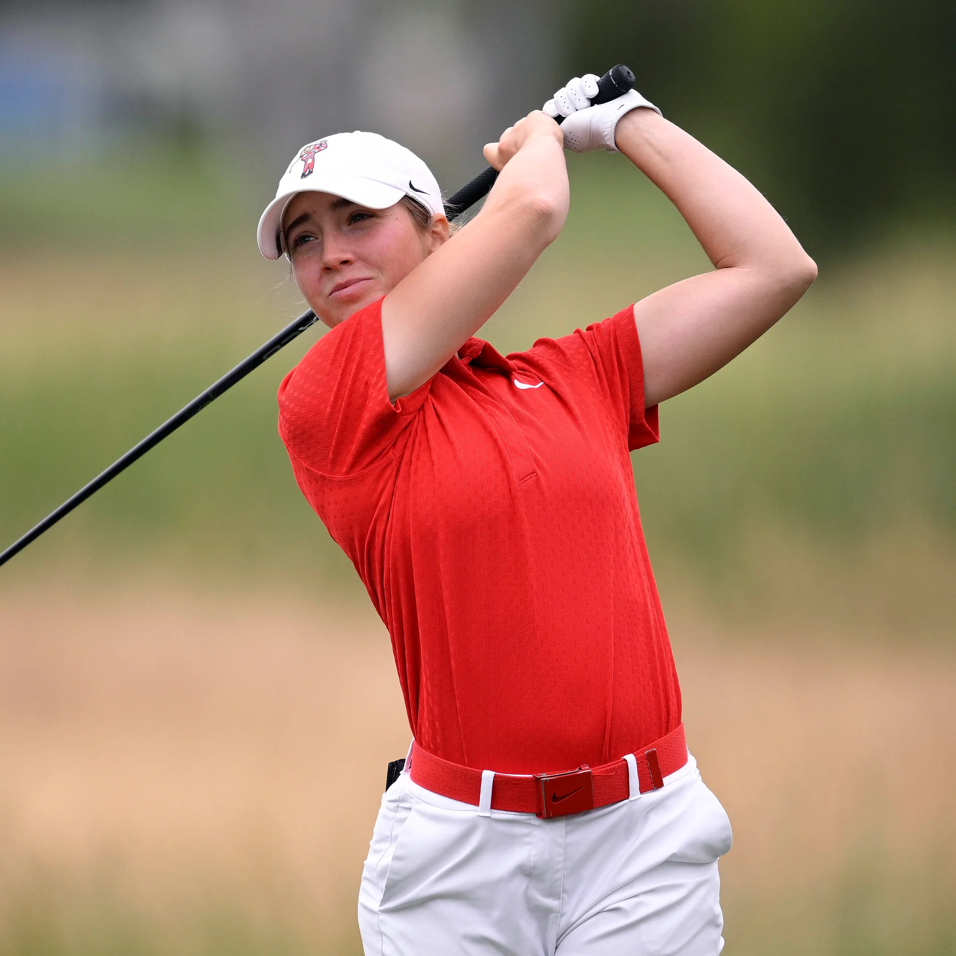 Golfer Kary Hollenbaugh follows through on a swing, wearing a white athletic cap, a bright red short-sleeve polo with a matching red belt, and white trousers.