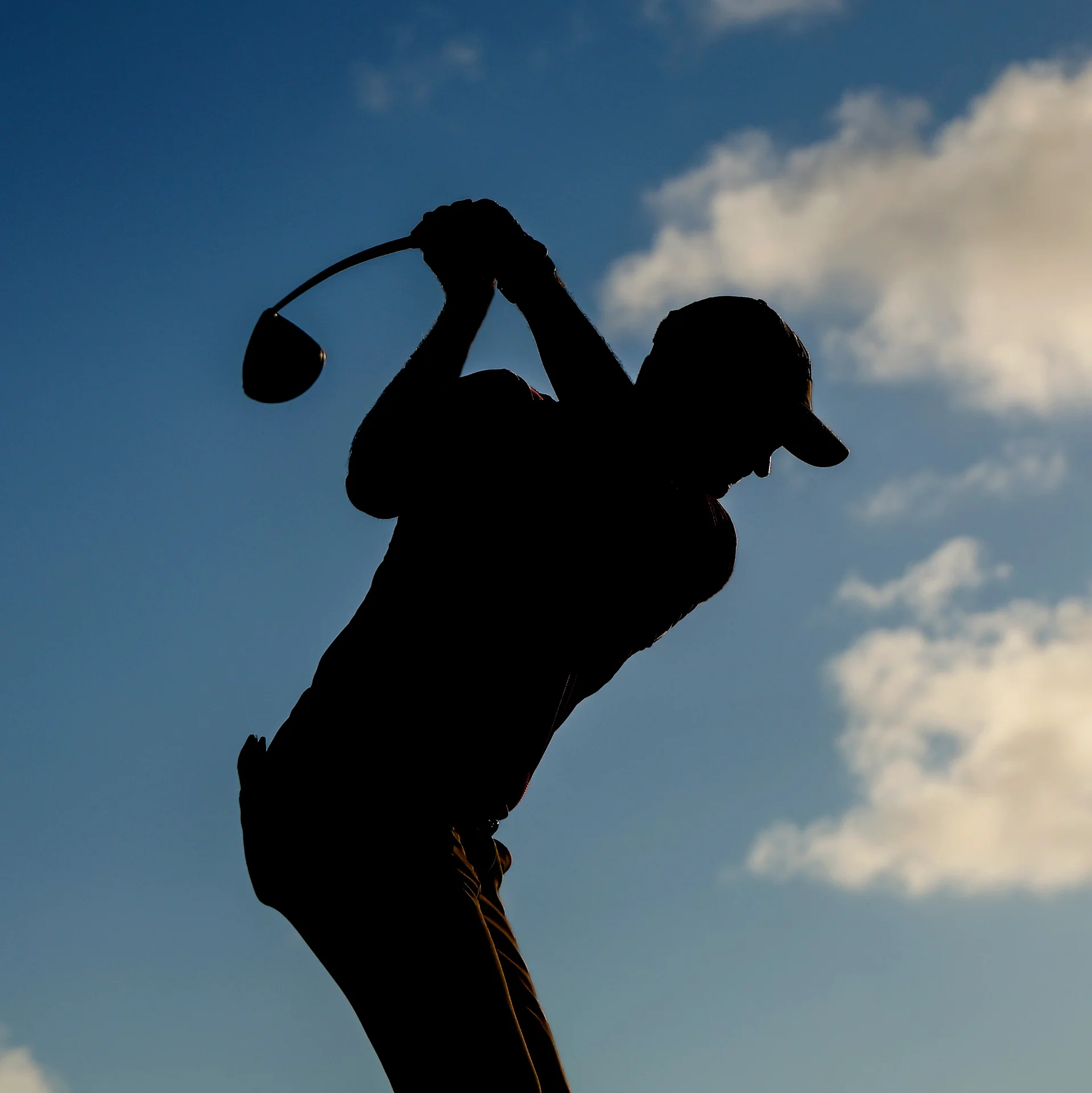 A silhouette of a male golfer teeing off with a driver set against blue sky.