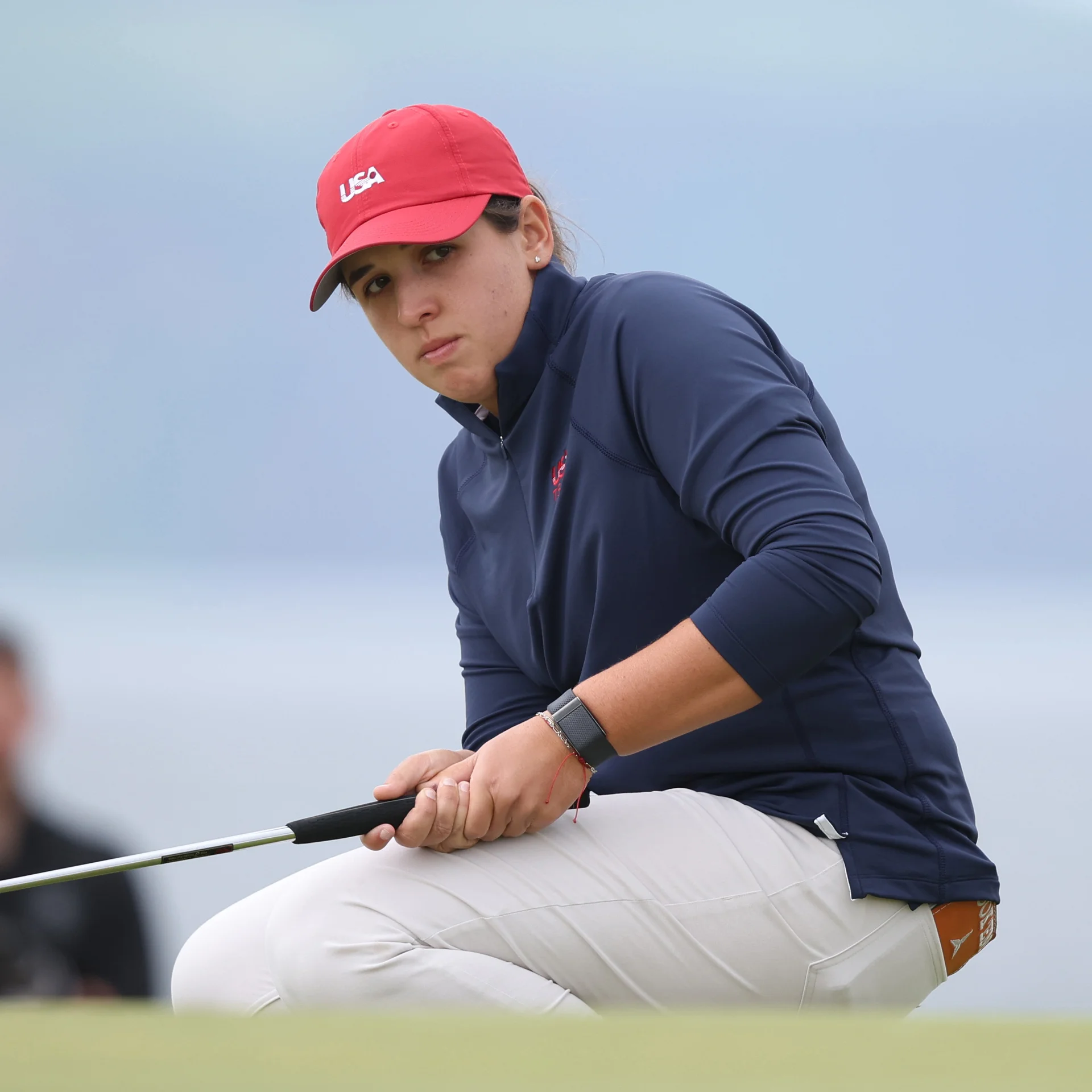 Golfer Farah O'Keefe crouches low on the green to read a putt, wearing a red USA cap, a navy blue long-sleeve pullover, and light-coloured trousers.