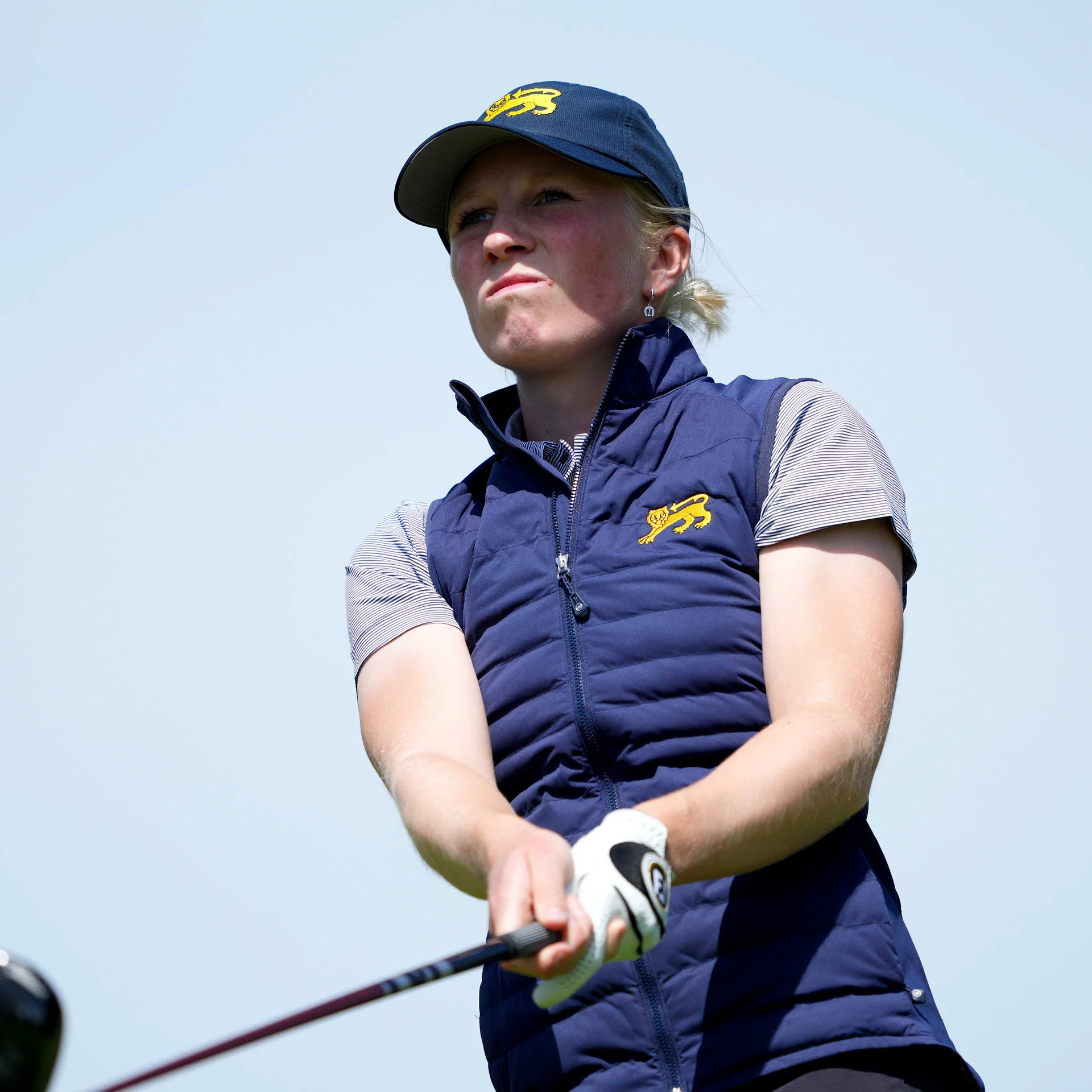 Golfer Nellie Ong, wearing a navy cap and matching vest with a yellow lion logo, looks off into the distance with a determined expression against a clear sky.