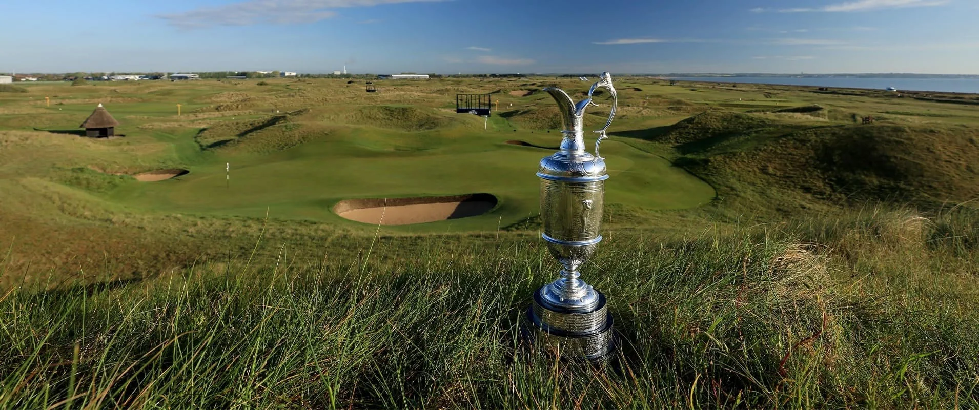 The Claret Jug sits in long grass with a golf green and links course in the background.