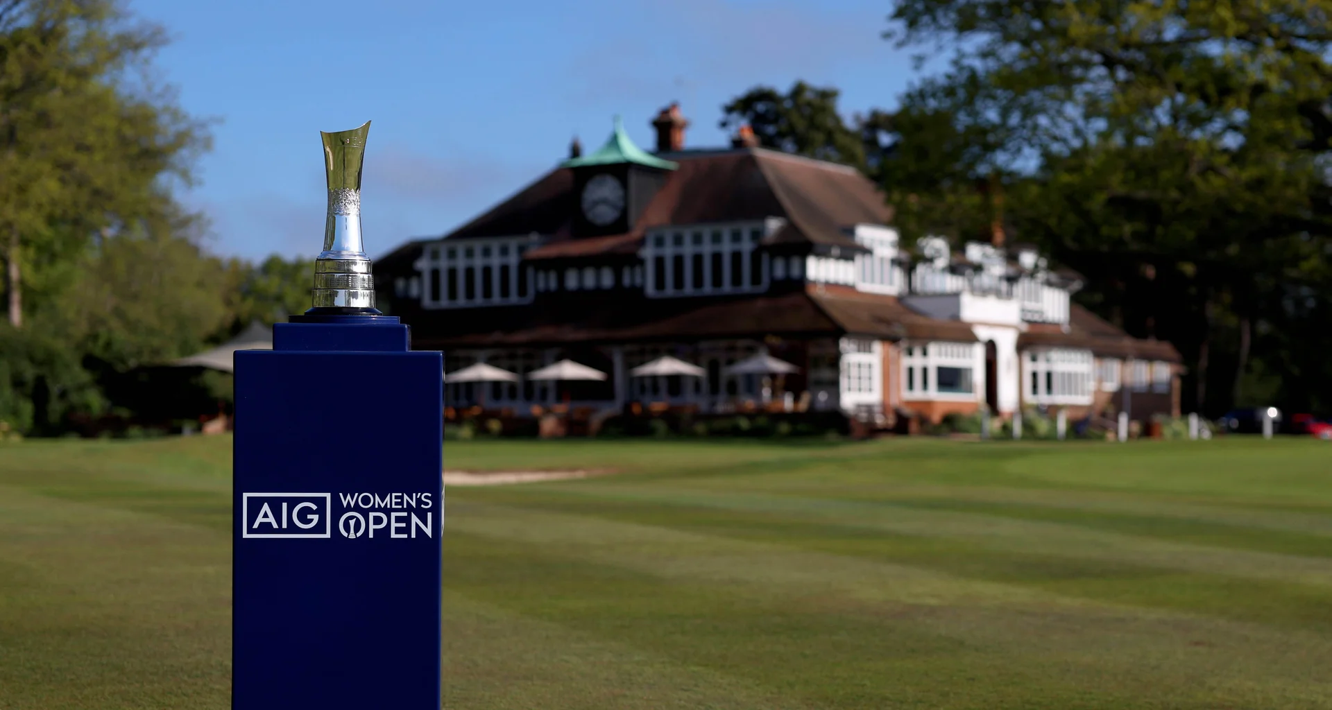 The AIG Women's Open Trophy sits on a blue branded podium in front of the clubhouse at Sunningdale.