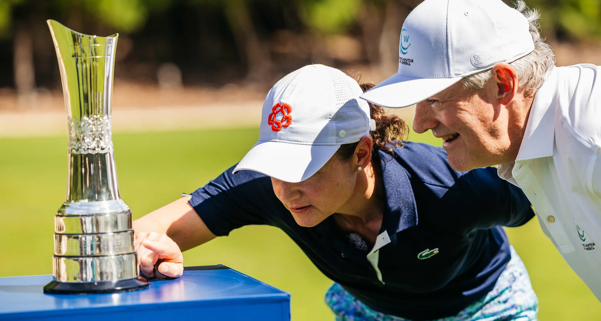 Lorena Ochoa points to her name on the AIG Women's Open trophy placed on a blue plinth. A man wearing a white cap looks to where she is pointing.