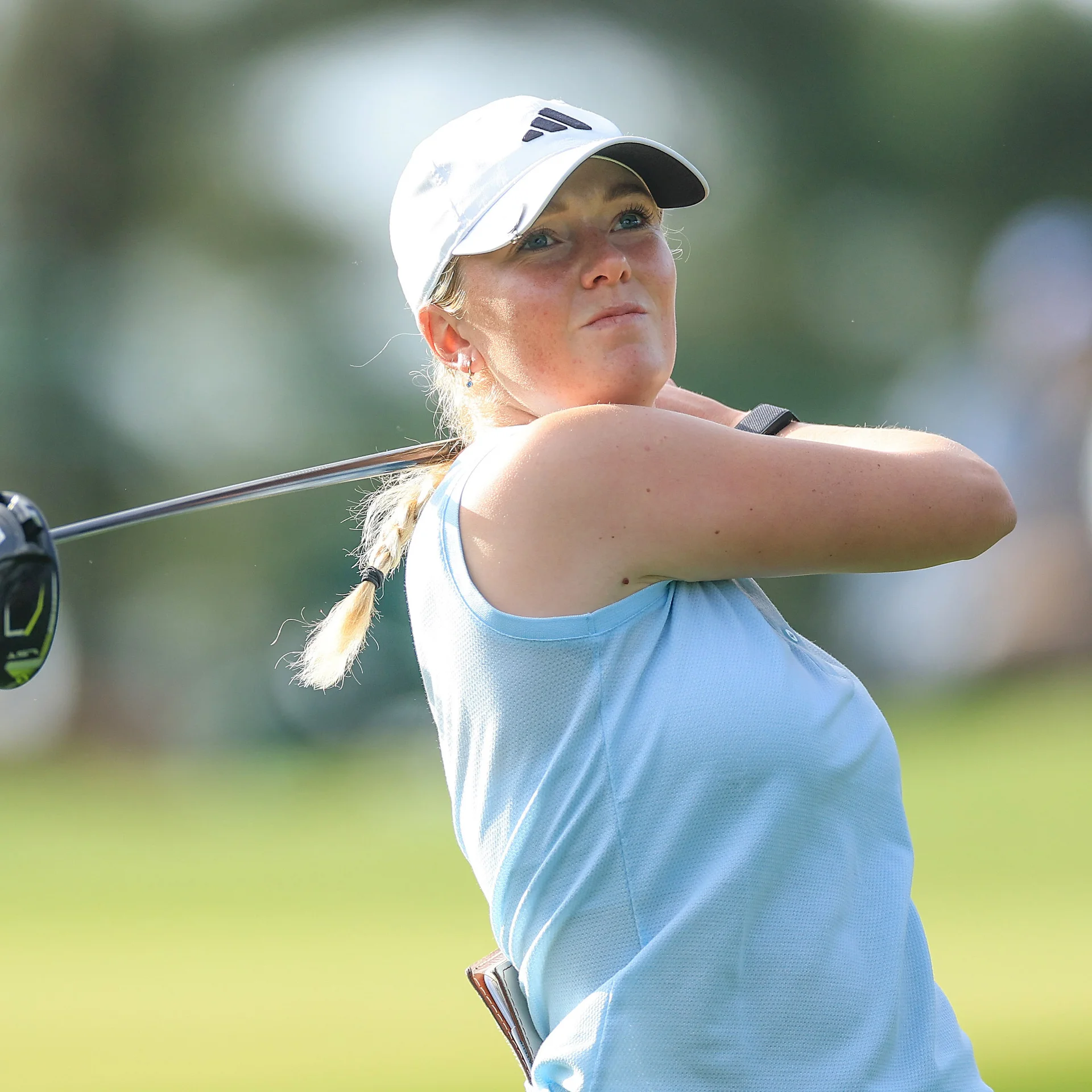 Golfer Patience Rhodes, wearing a white Adidas cap and light blue top, looks upward with a thoughtful expression following her swing.