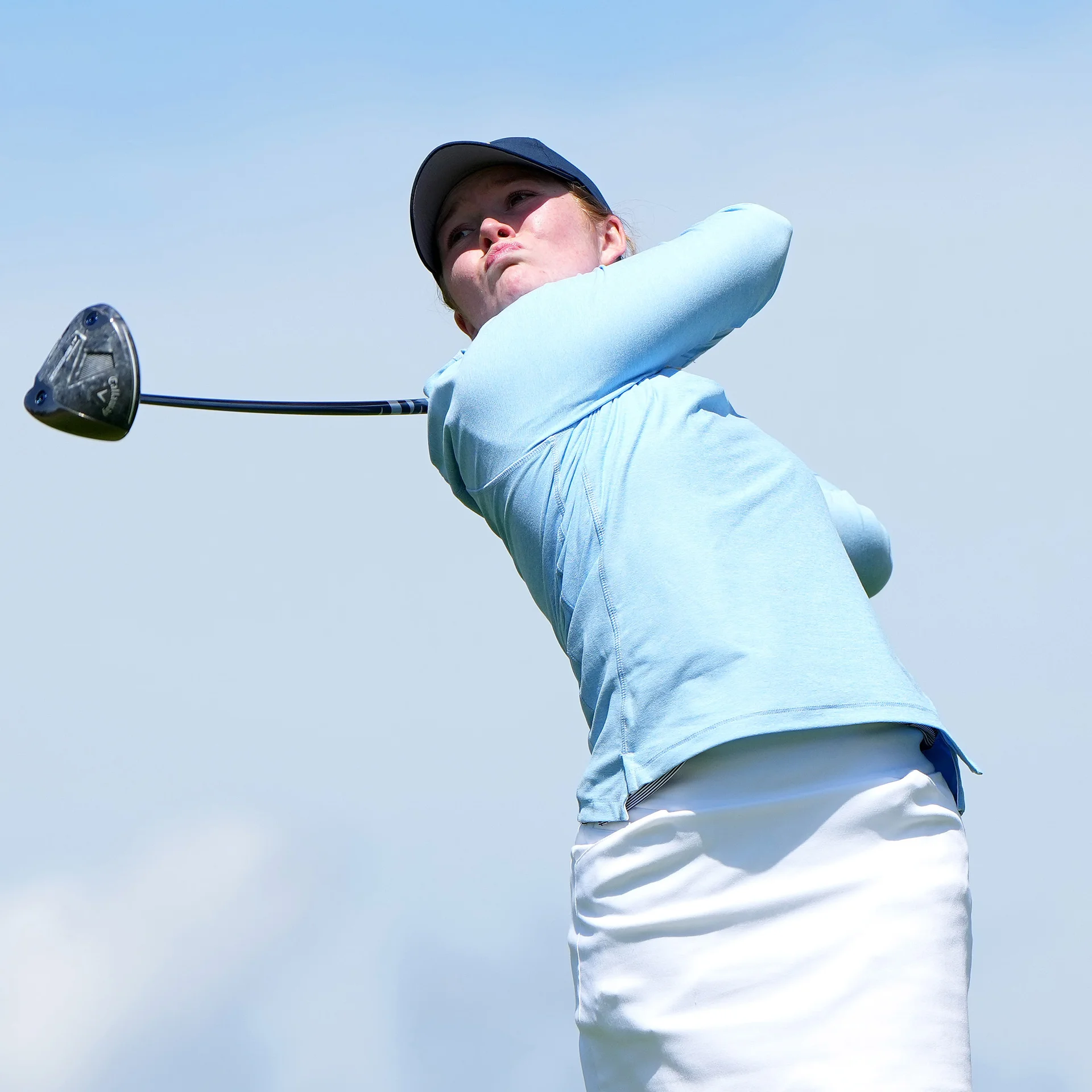 Golfer Isla McDonald-O'Brien tracks her ball flight during a powerful swing, wearing a light blue long-sleeve shirt and a dark blue cap against a bright sky.