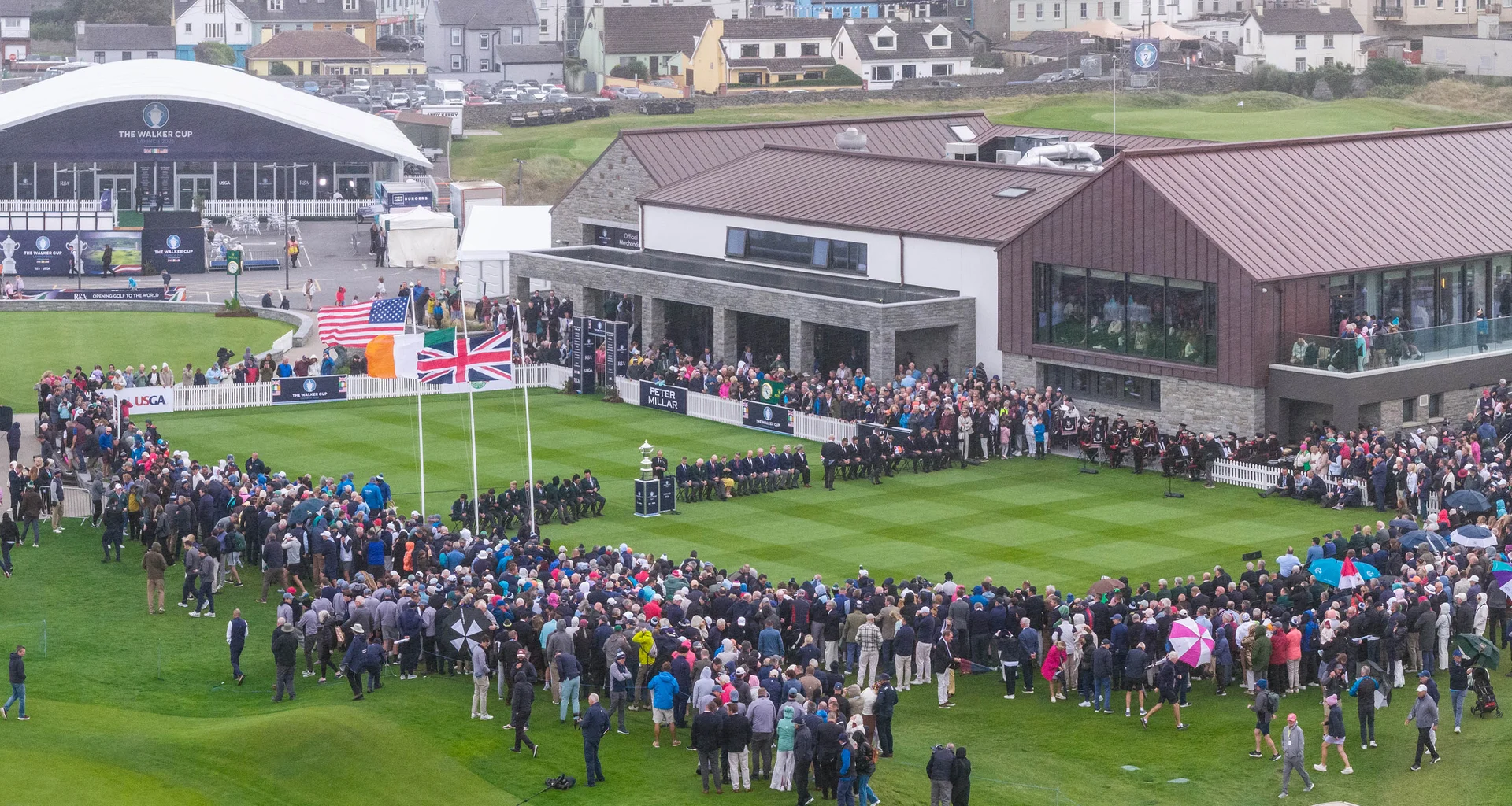 A crowd is gathered around the first tee at Lahinch for the opening ceremony of the Walker Cup. The flags Great Britain, Ireland and the USA are all raised on flagpoles and are blowing in the wind.