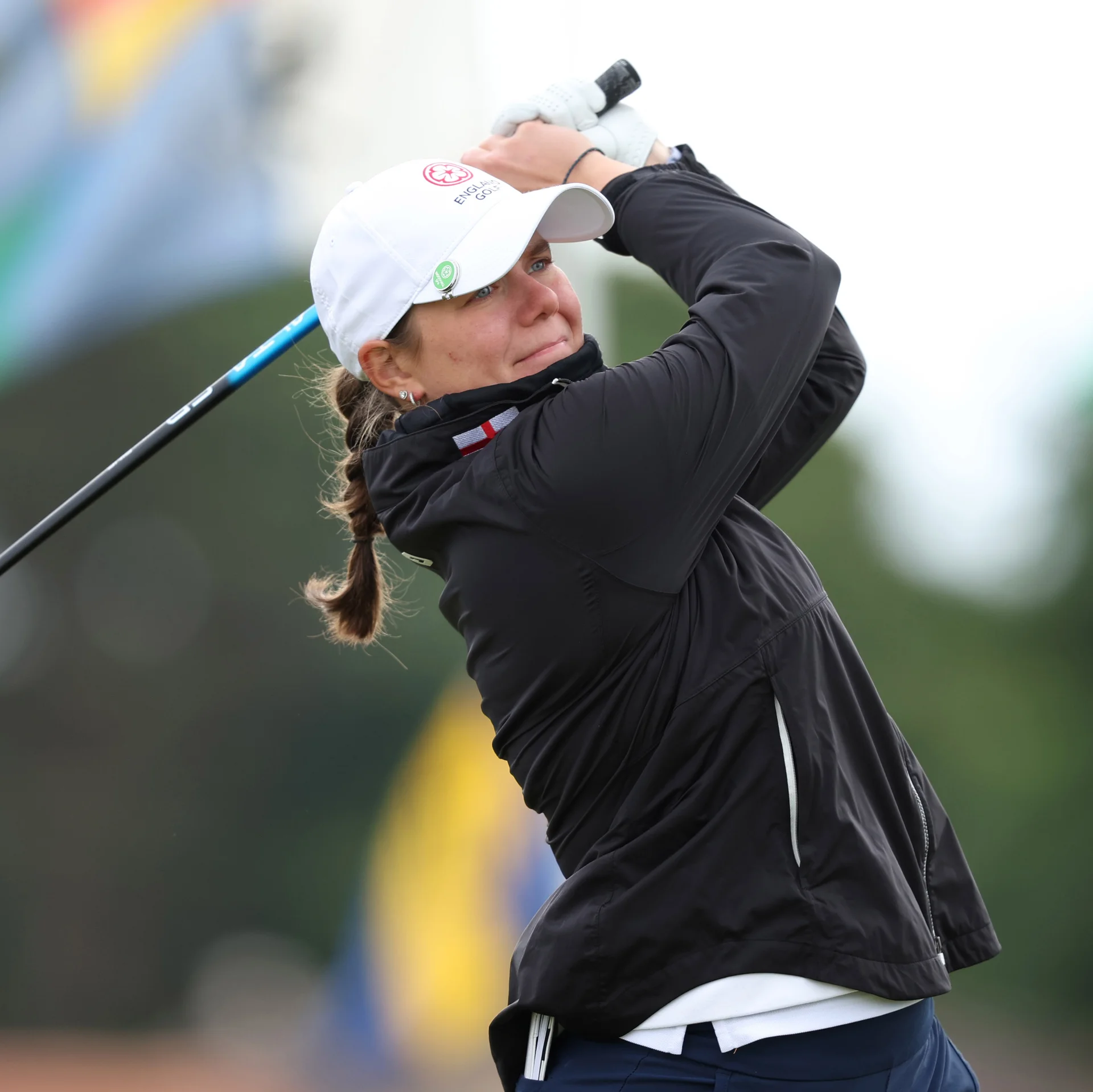 Golfer Lily Hirst smiles slightly at the top of her swing, wearing a white England Golf cap and a black windbreaker jacket.