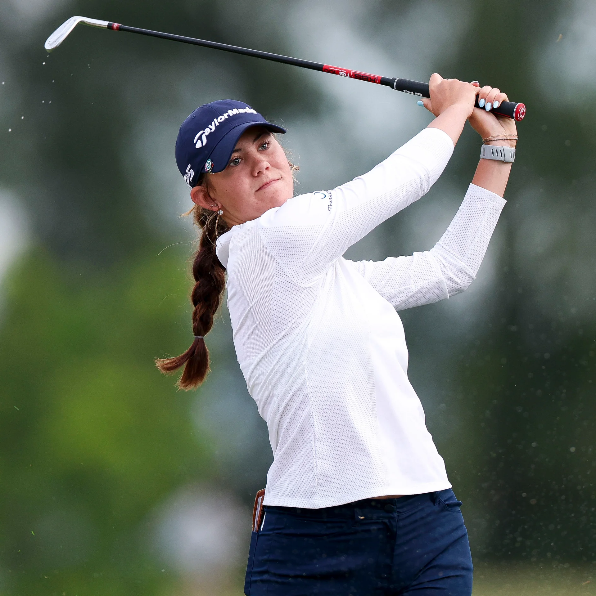 Golfer Asterisk Talley executes a powerful swing on the course, wearing a navy blue TaylorMade cap, a white long-sleeve athletic top, and dark trousers.