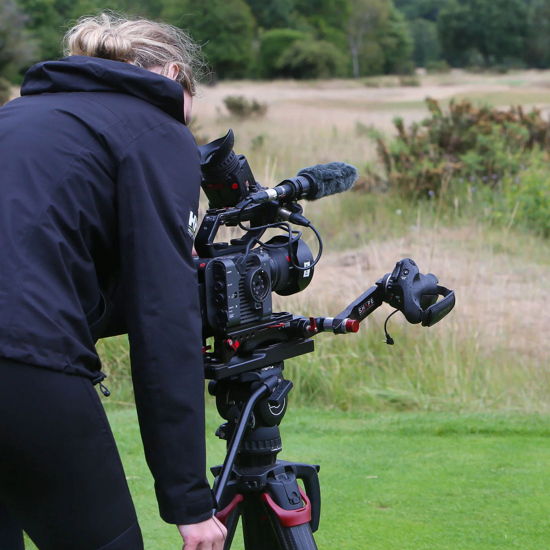 A camera operator looks down their lens towards some bushes on a golf course.