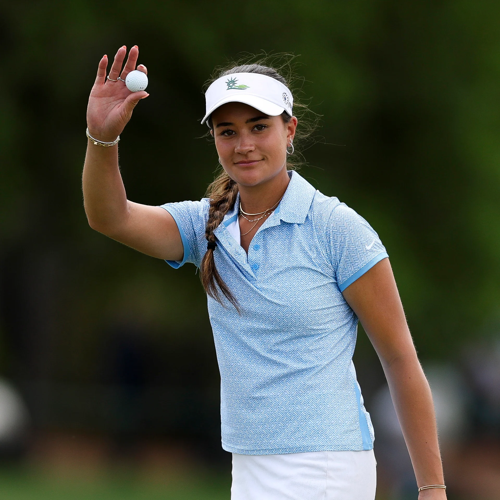 Golfer Kiara Romero smiles and holds up a golf ball in her right hand, wearing a white visor, a light blue patterned polo shirt, and a white skirt.