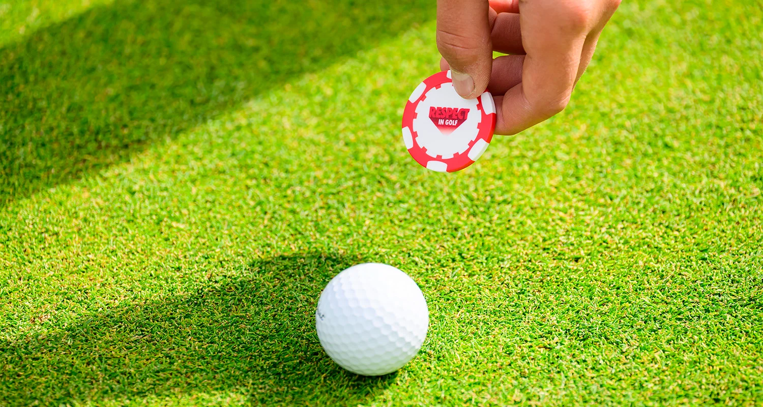 A hand holds a ball marker, which reads 'Respect in Golf', towards a golf ball on a green.