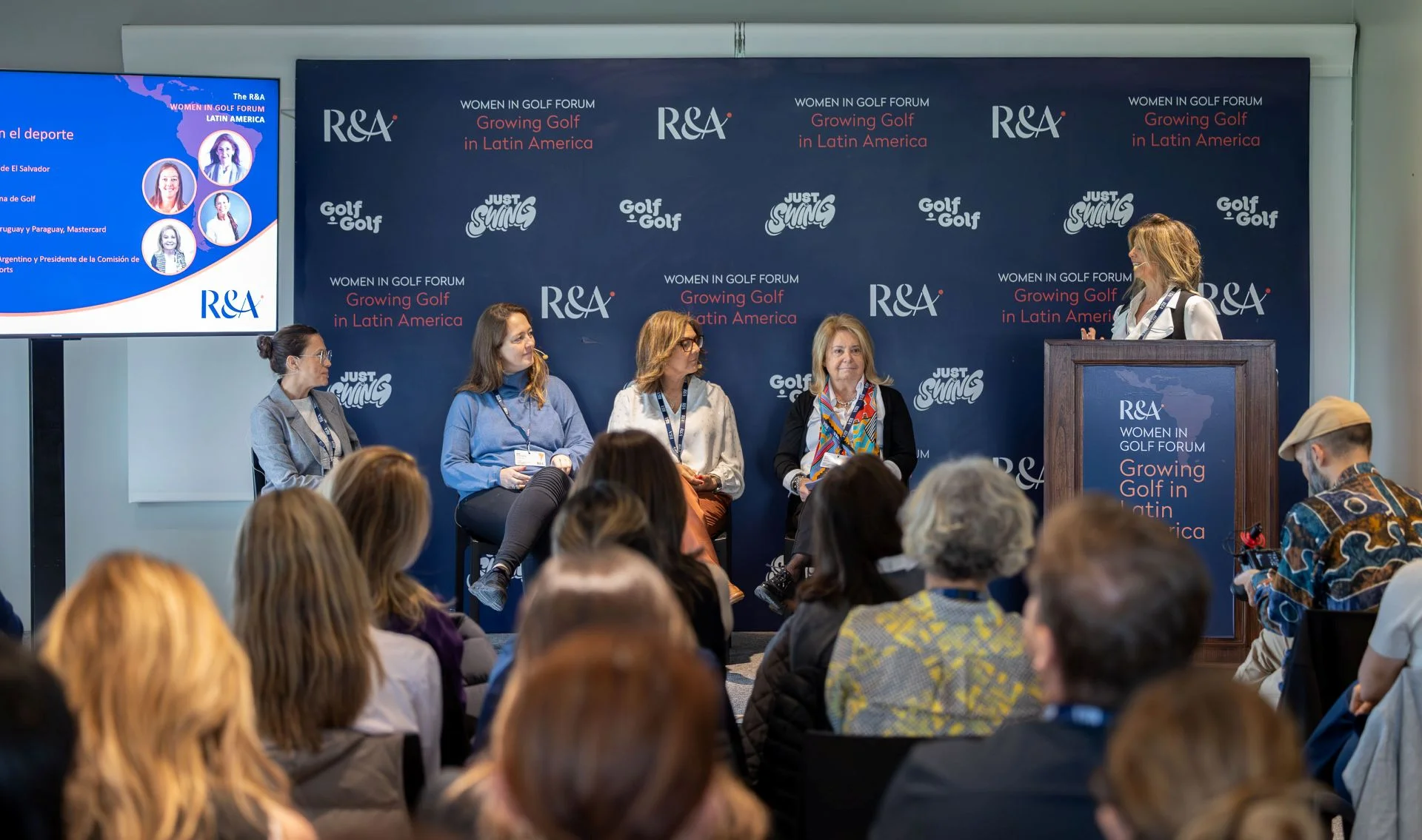 Four women sit and one stands at a podium on a raised stage in front of an audience.