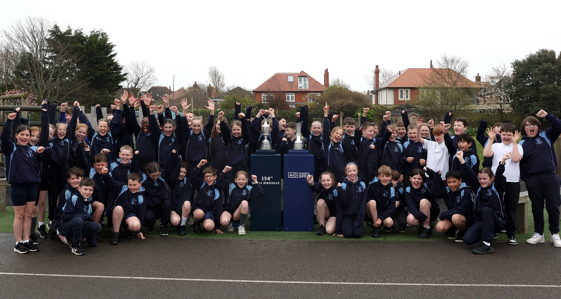 School children gather around The Claret Jug and AIG Women's Open Trophy in a school playground.