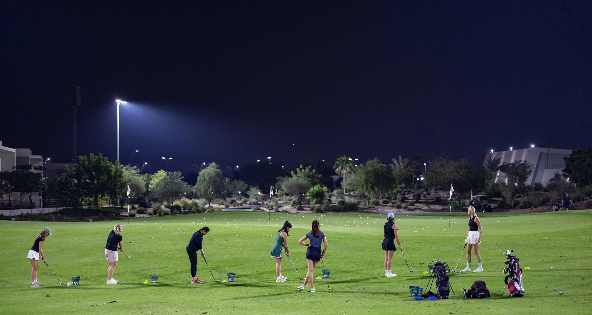 A group of golfers practicing around a green, at a floodlit practice area.