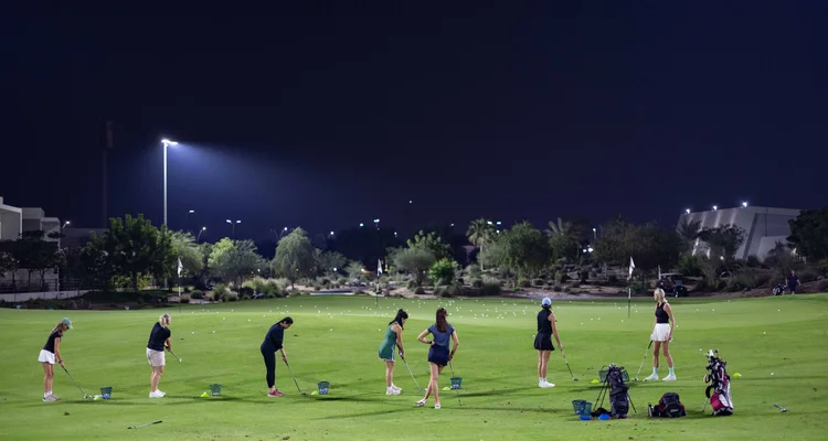 A group of golfers practicing around a green, at a floodlit practice area.