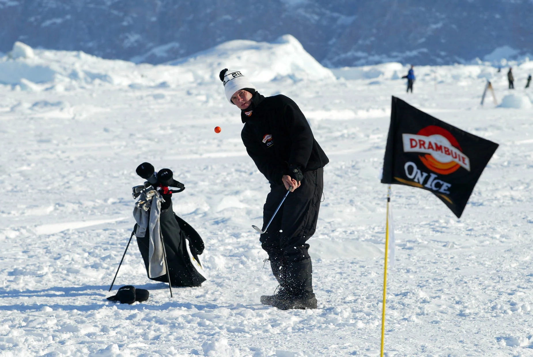 Golfer chips an orange golf ball over ice and snow towards a golf hole.