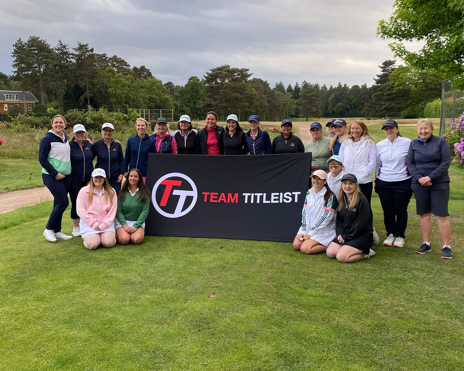 A group of woman stand behind and kneel beside a banner which reads Team Titleist on the tee area of a golf course.