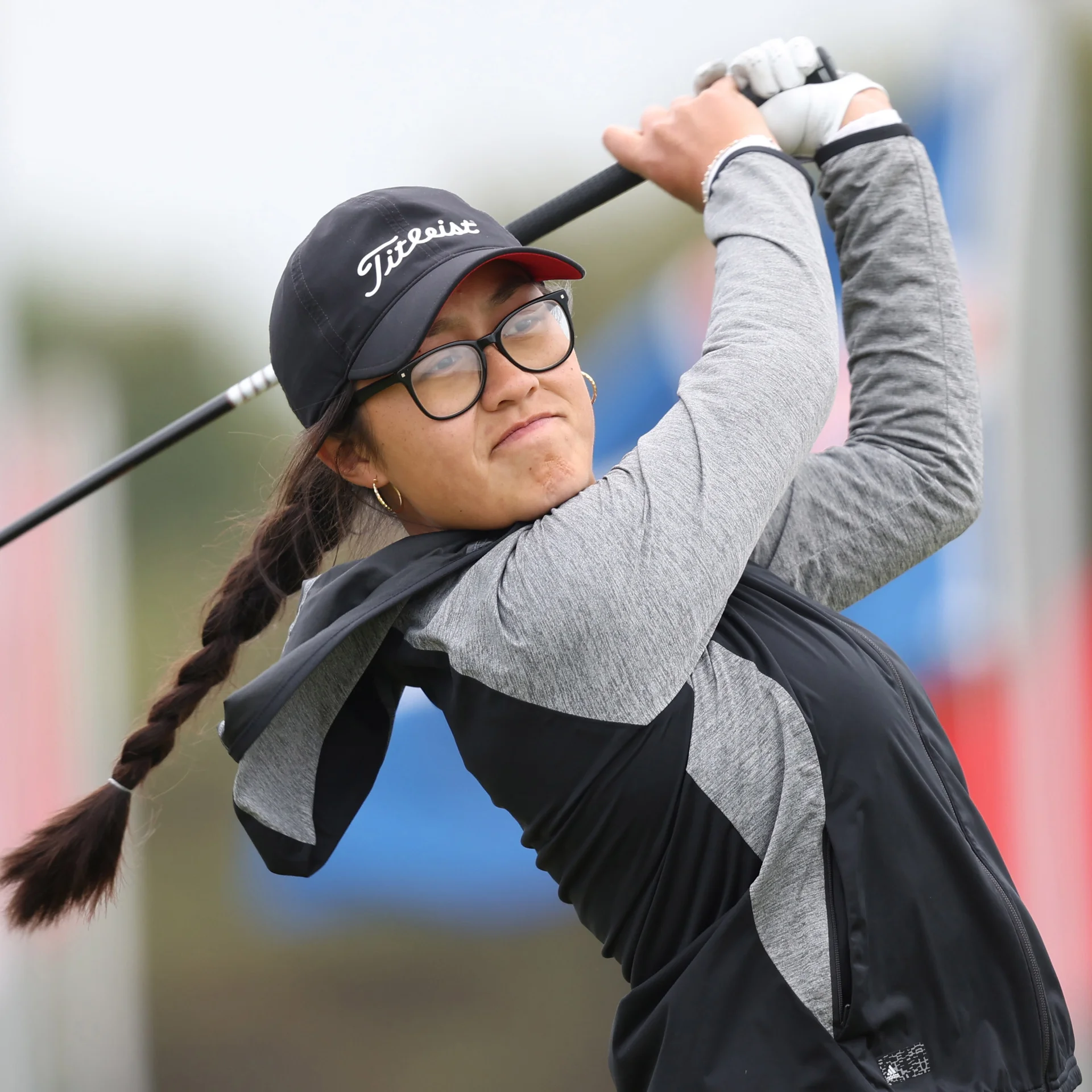 Golfer Davina Xanh, wearing a black Titleist cap and glasses, is captured mid-swing looking focused with her long dark braid over her shoulder.