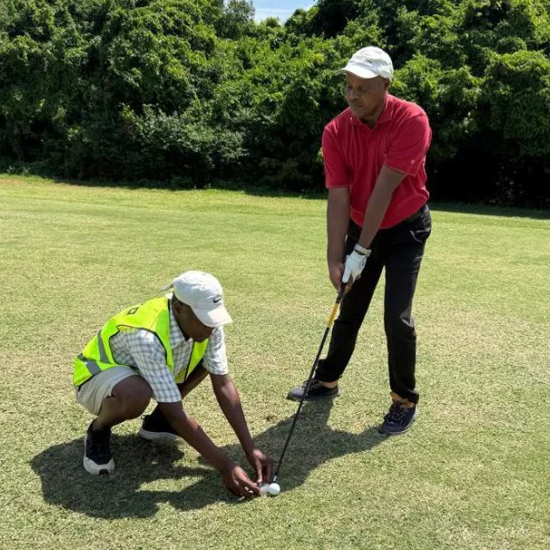 A visually-impaired golfer in a red t-shirt and white cap stands at a tee ready to hit his golf shot. A guide is crouched down by his club lining up the golfer's shot.