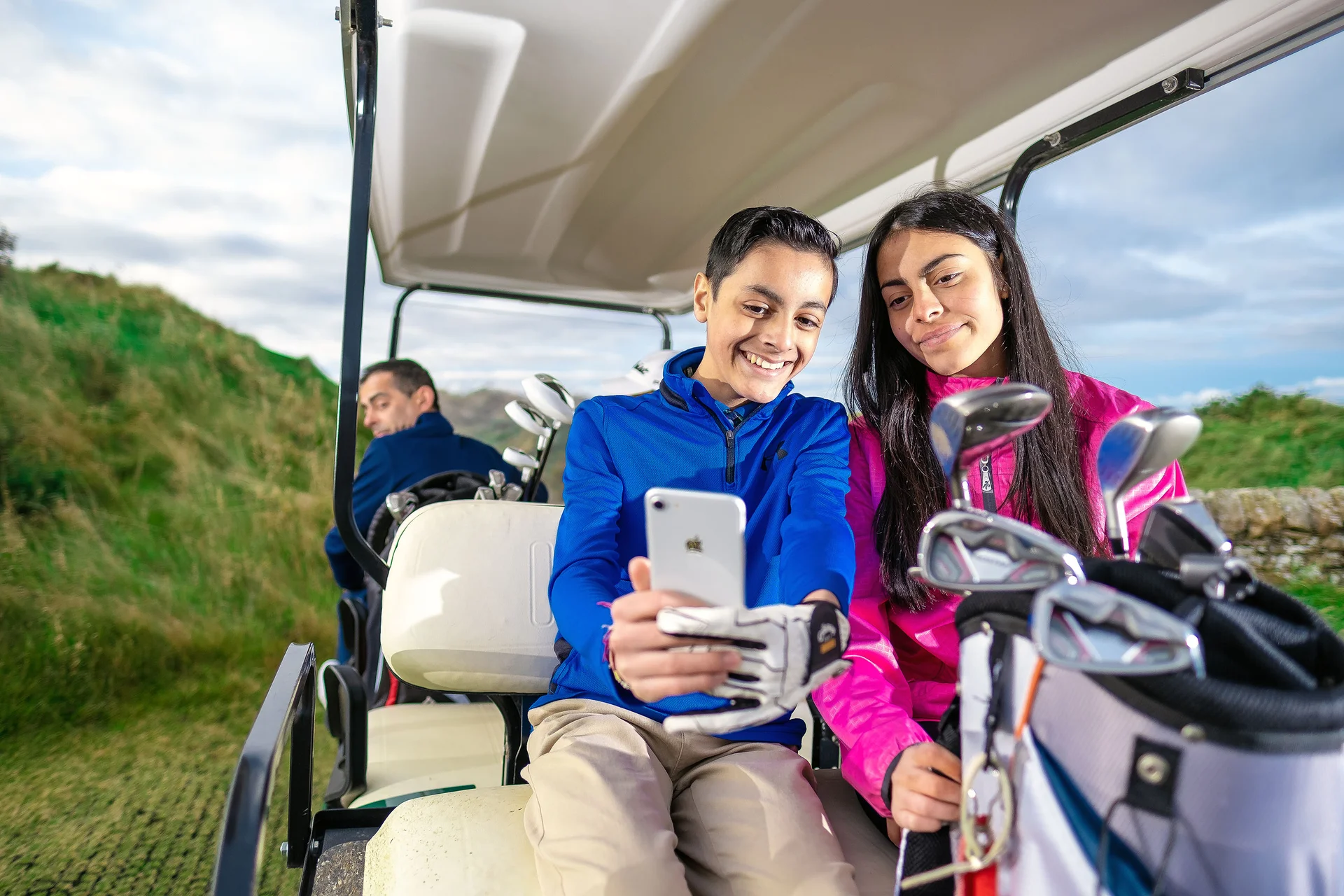 Two junior golfers take a selfie in the back of a golf cart as an adult, sitting in the front looks back at them.