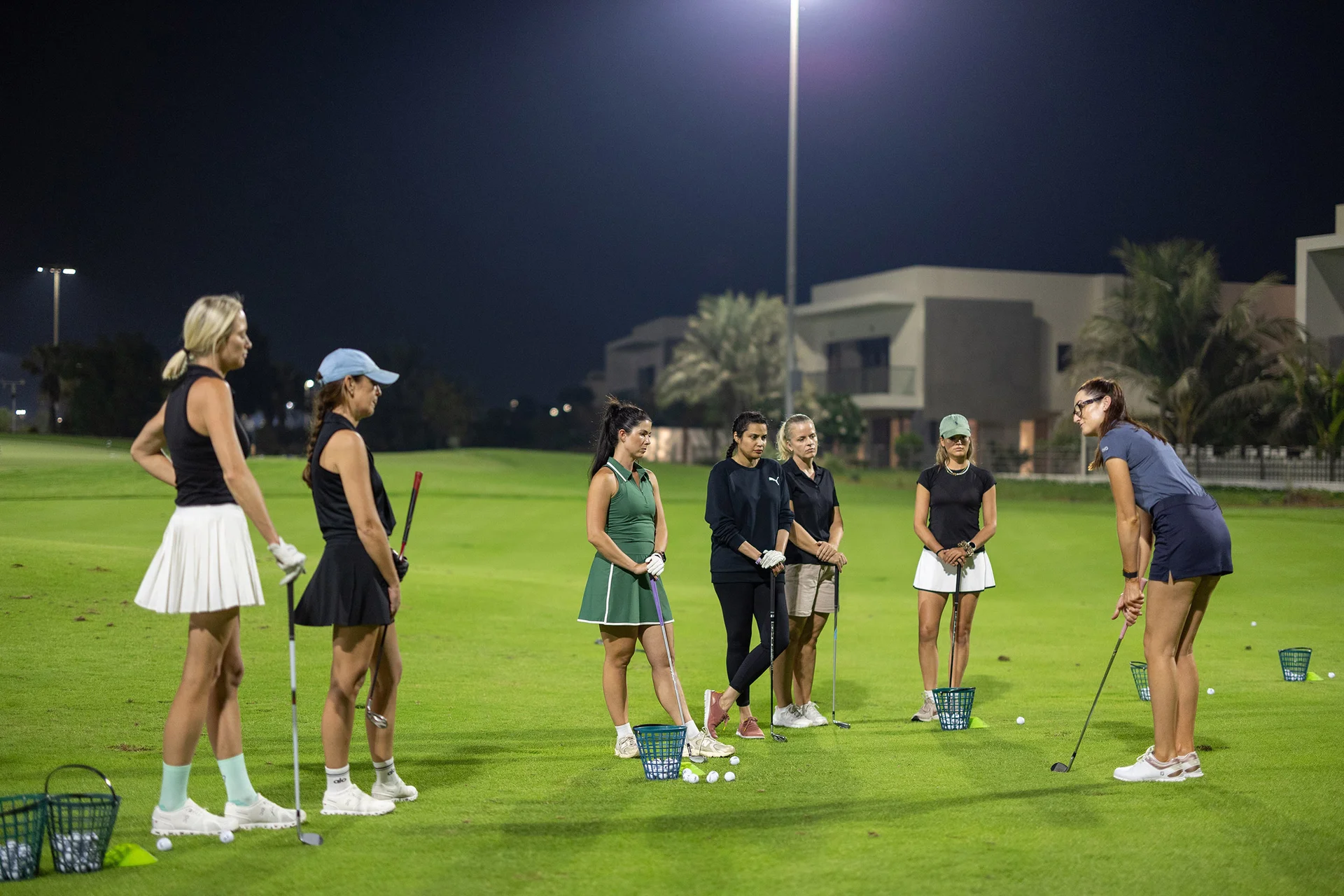 A group of women stand listening to a female golf instructor at a driving range at night under floodlights.