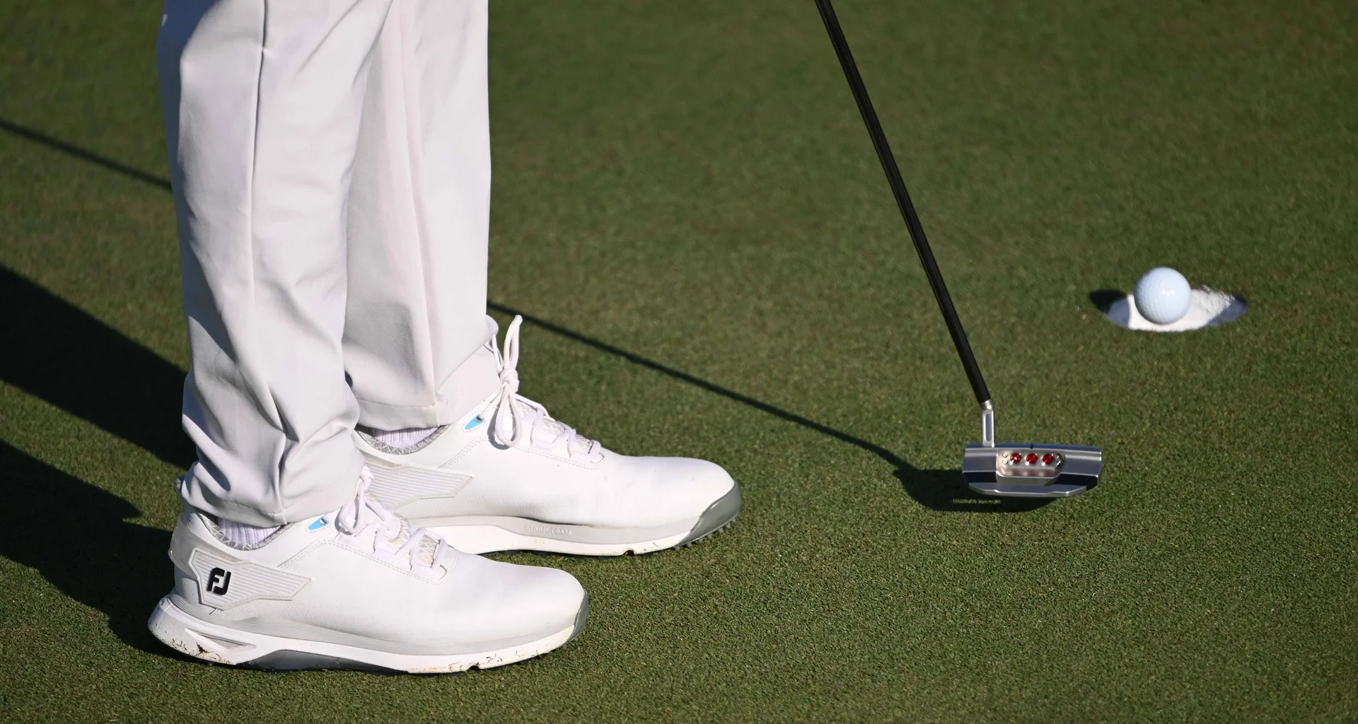 A golf ball drops in to the hole with a putter and two legs in shot.