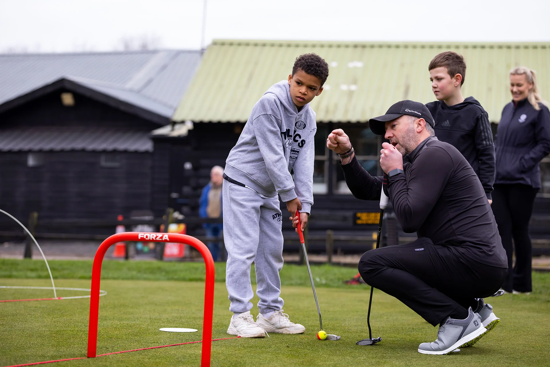 A golf professional kneels down and gives golfing tips to a child, aged around 10 years old, who is lining up a putt.