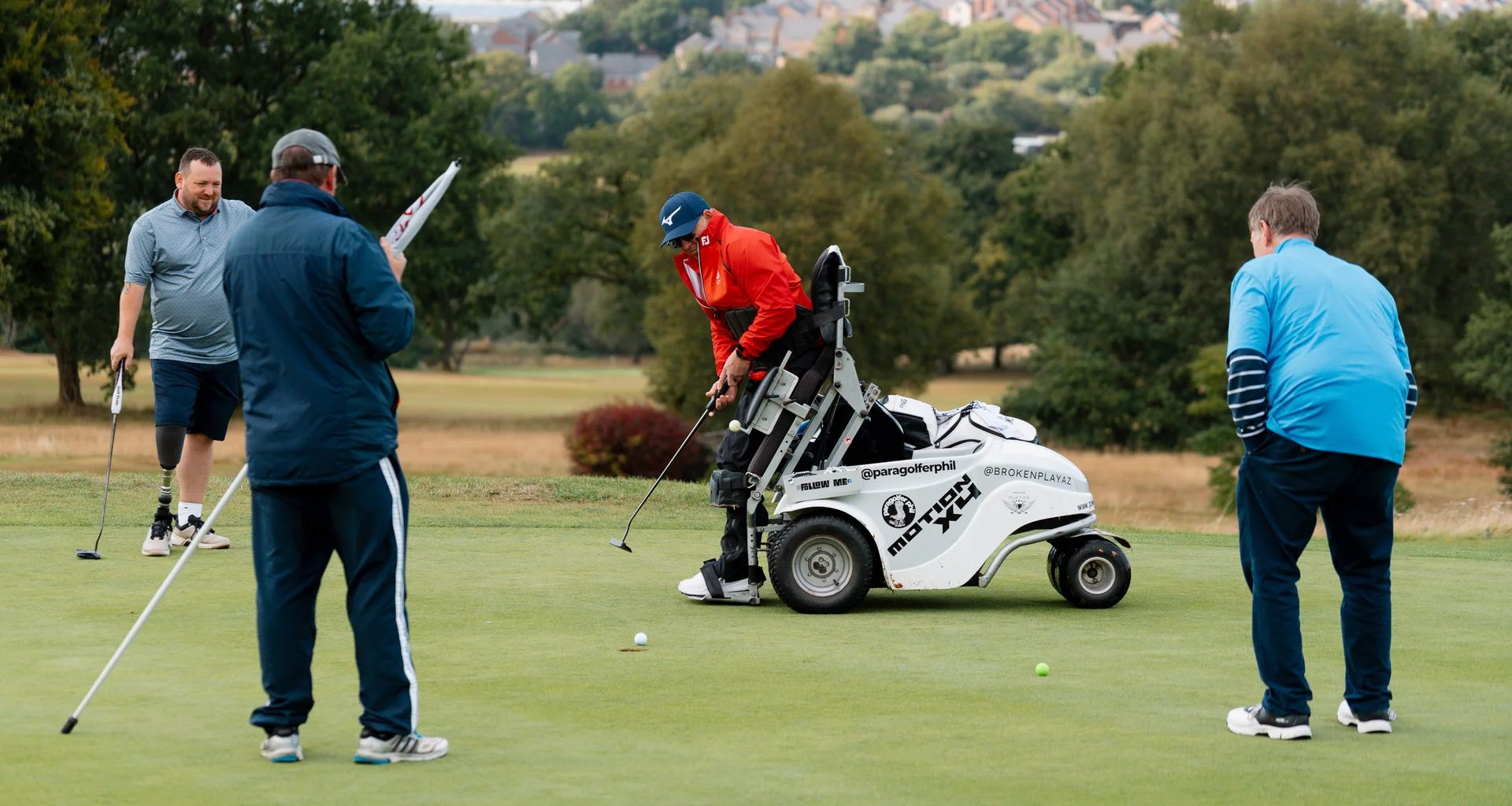 Four golfers on a green. One golfer, using a mobility chair, putts the ball towards the hole as the others watch on.