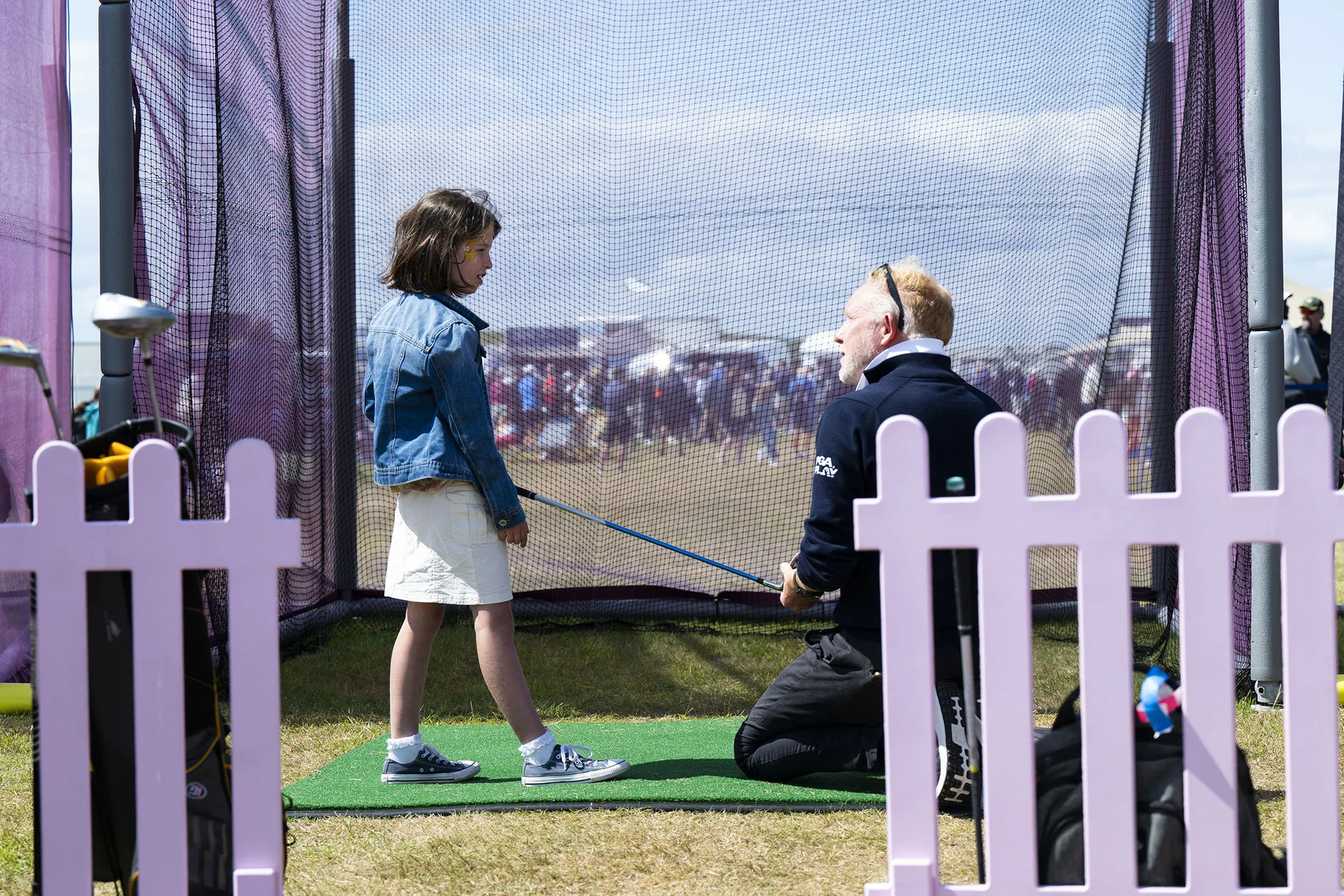 A young girls listens as she is given a golfing lesson in front of a net. Behind the net there is a festival atmosphere and stalls in the distance.