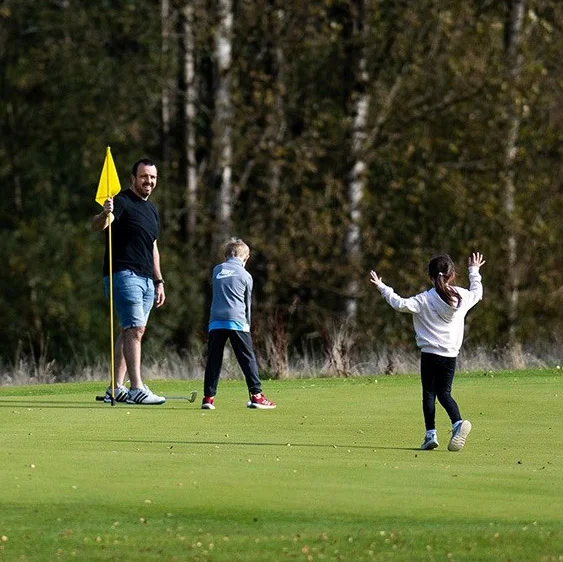 A man and a young boy and girl stand happily on a golf green. The man hold the pin flag upright while the boy plays a shot.