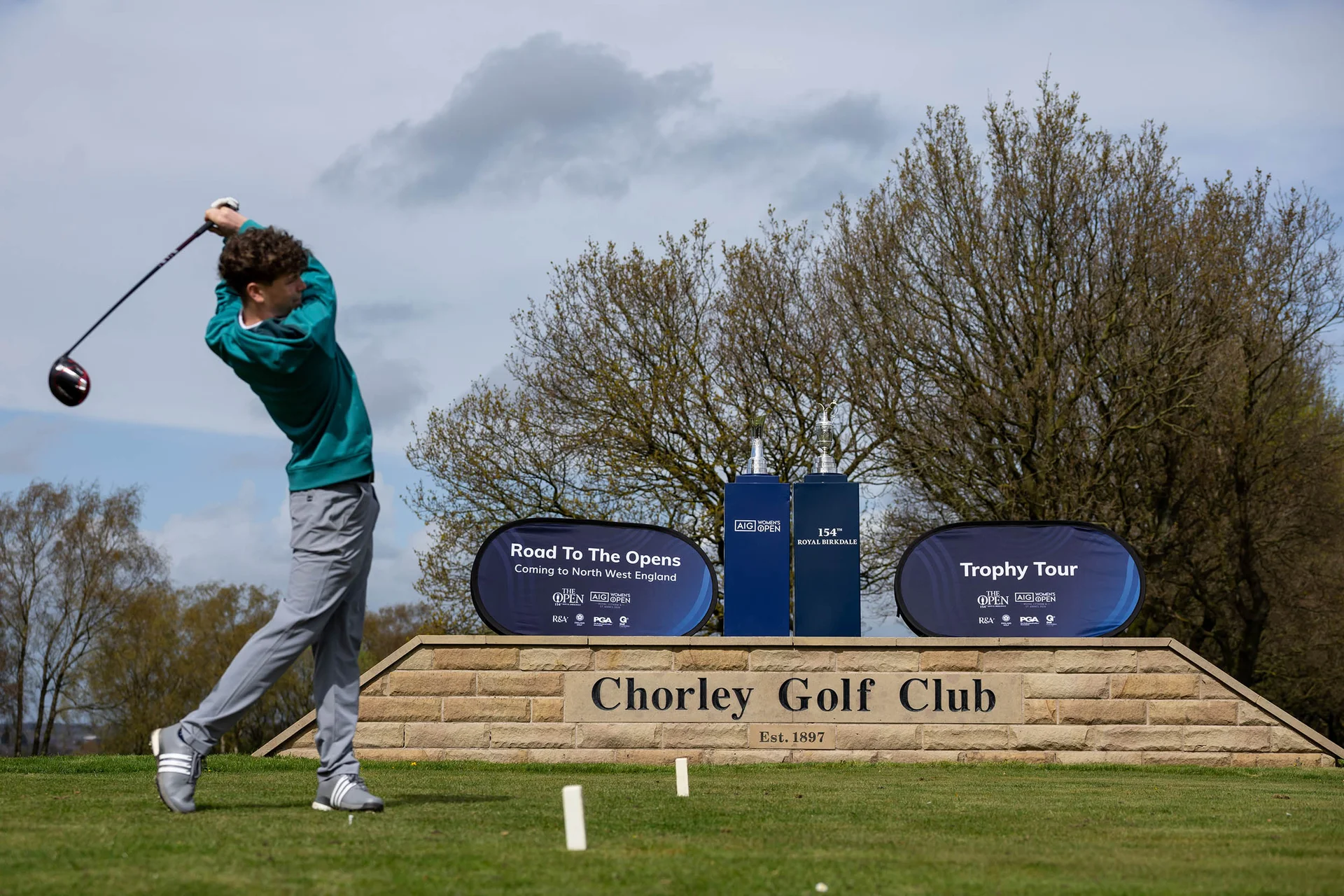 A golfer completes a swing after teeing off infront of The Claret Jug and AIG Women's Open trophy and sign which reads Chorley Golf Club.