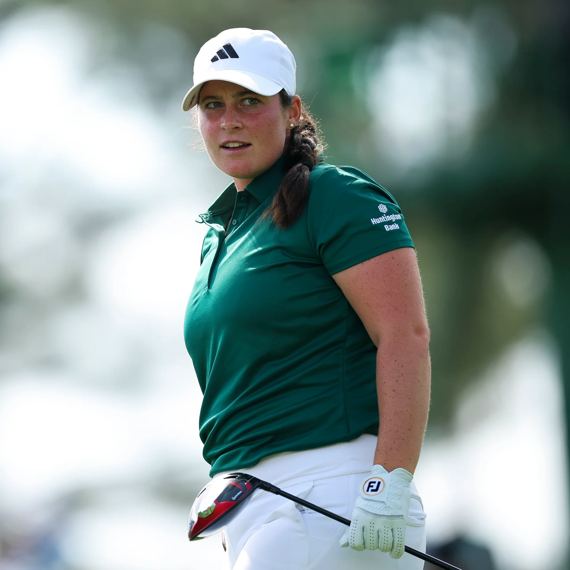 Golfer Avery Weed looks over her shoulder with a focused expression, wearing a white Adidas cap, a dark green polo shirt with a Huntington Bank logo, and white trousers.
