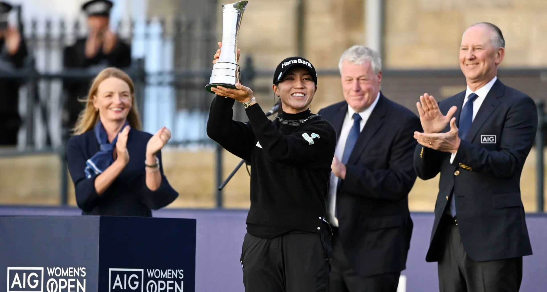 Lydia Ko, the 2024 AIG Women’s Open champion, holds her trophy as Peter Zaffino, CEO and Chairman of AIG, and others celebrate her win in the background.