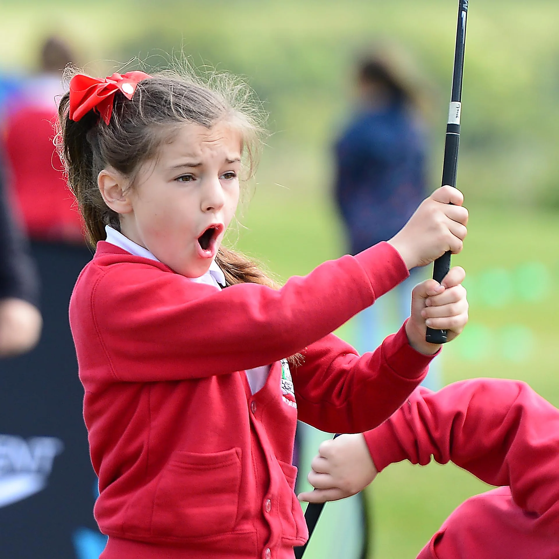 A young girl wearing a red school cardigan looks on after hitting a golf shot.