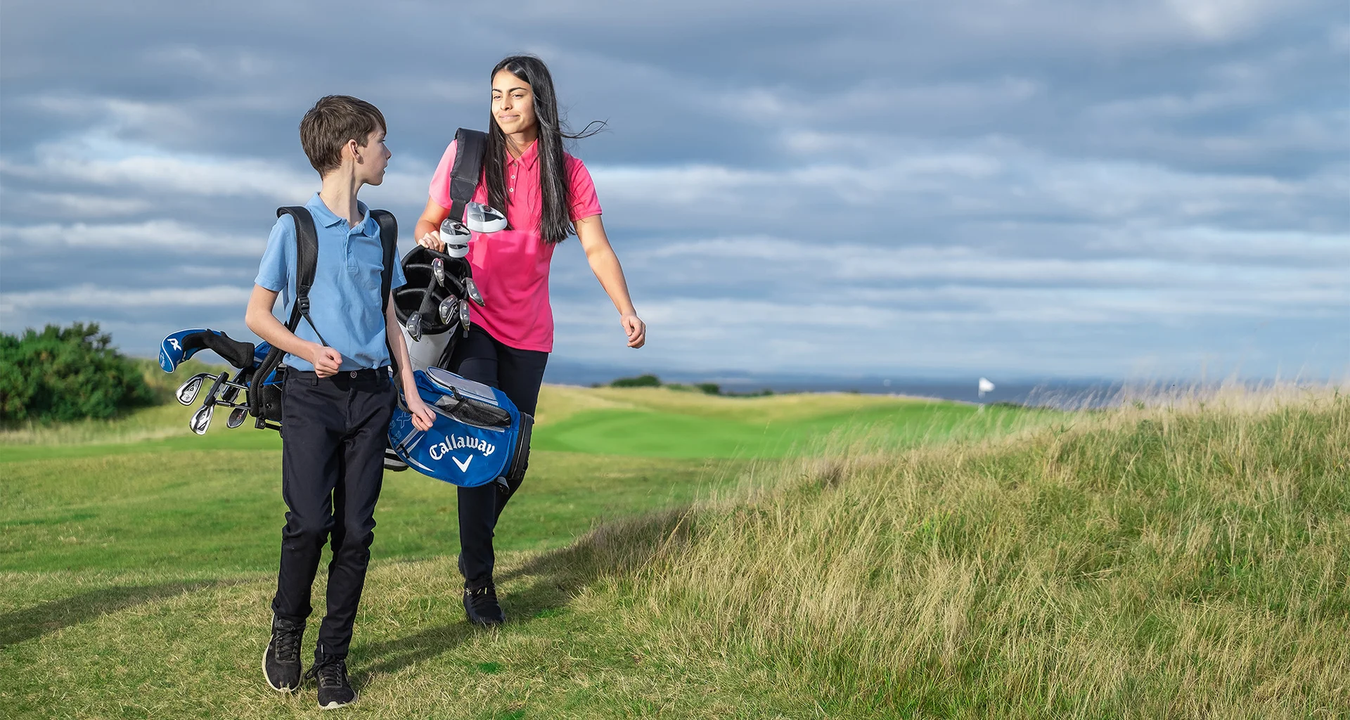 Two young people, a boy and girl in their early teens, look at each other smiling as they carry their golf bags on a golf course. 
