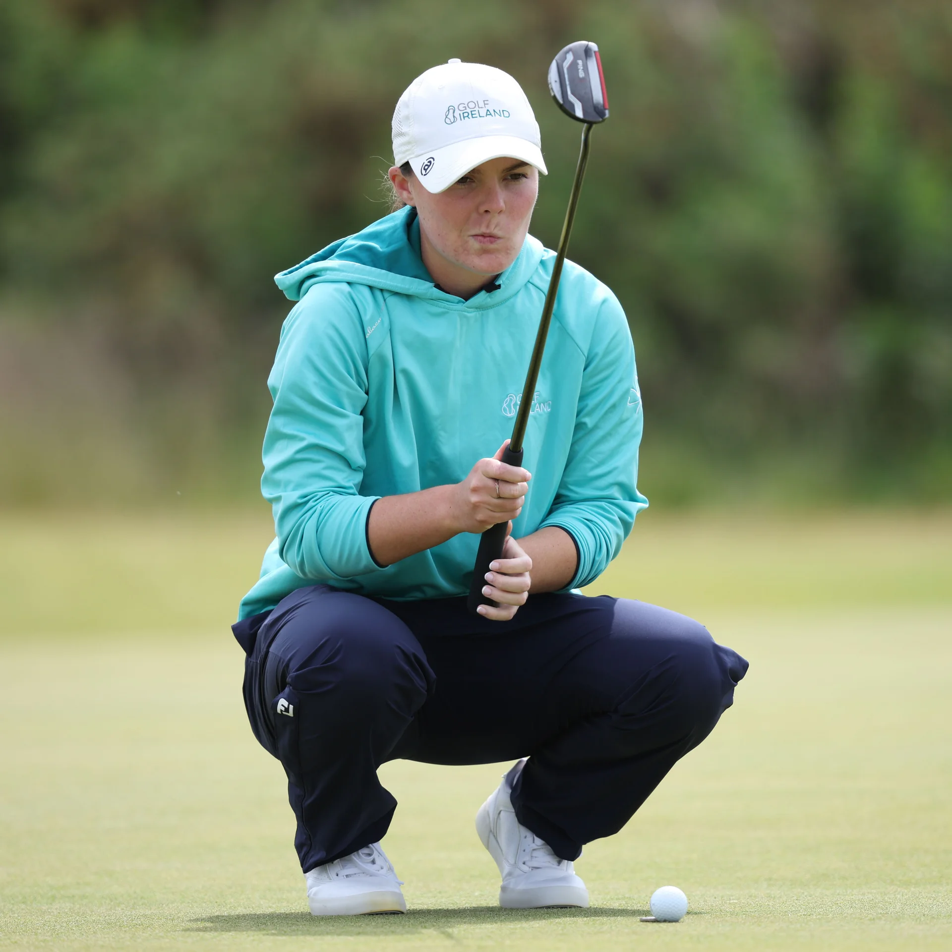 Golfer Beth Coulter, wearing a white Golf Ireland cap and a teal hoodie, crouches on the green to line up her putt.