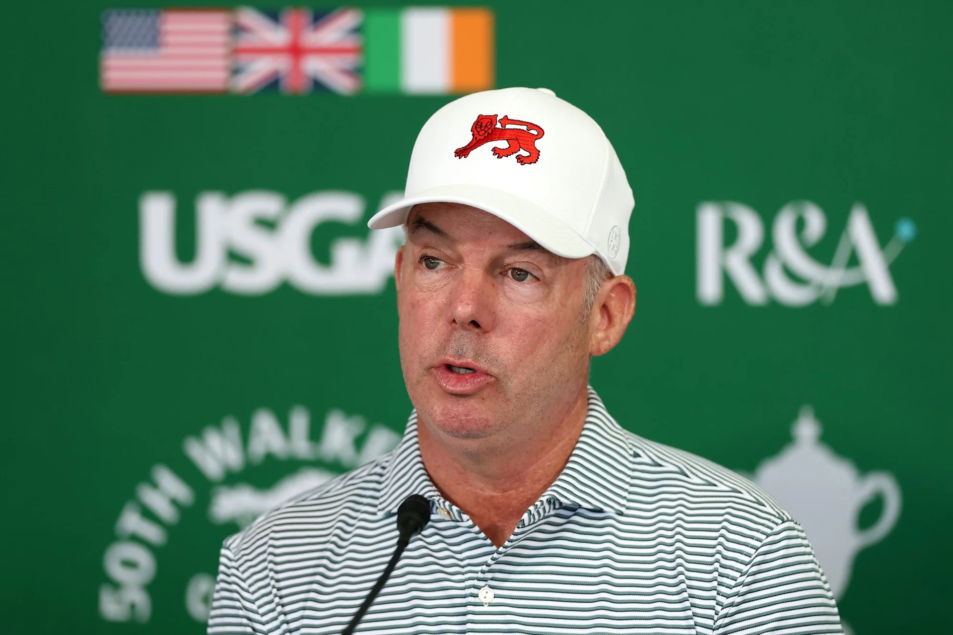 Dean Robertson speaks at a press conference in front of a green backdrop wit the USA, Great Britain and Ireland flags.