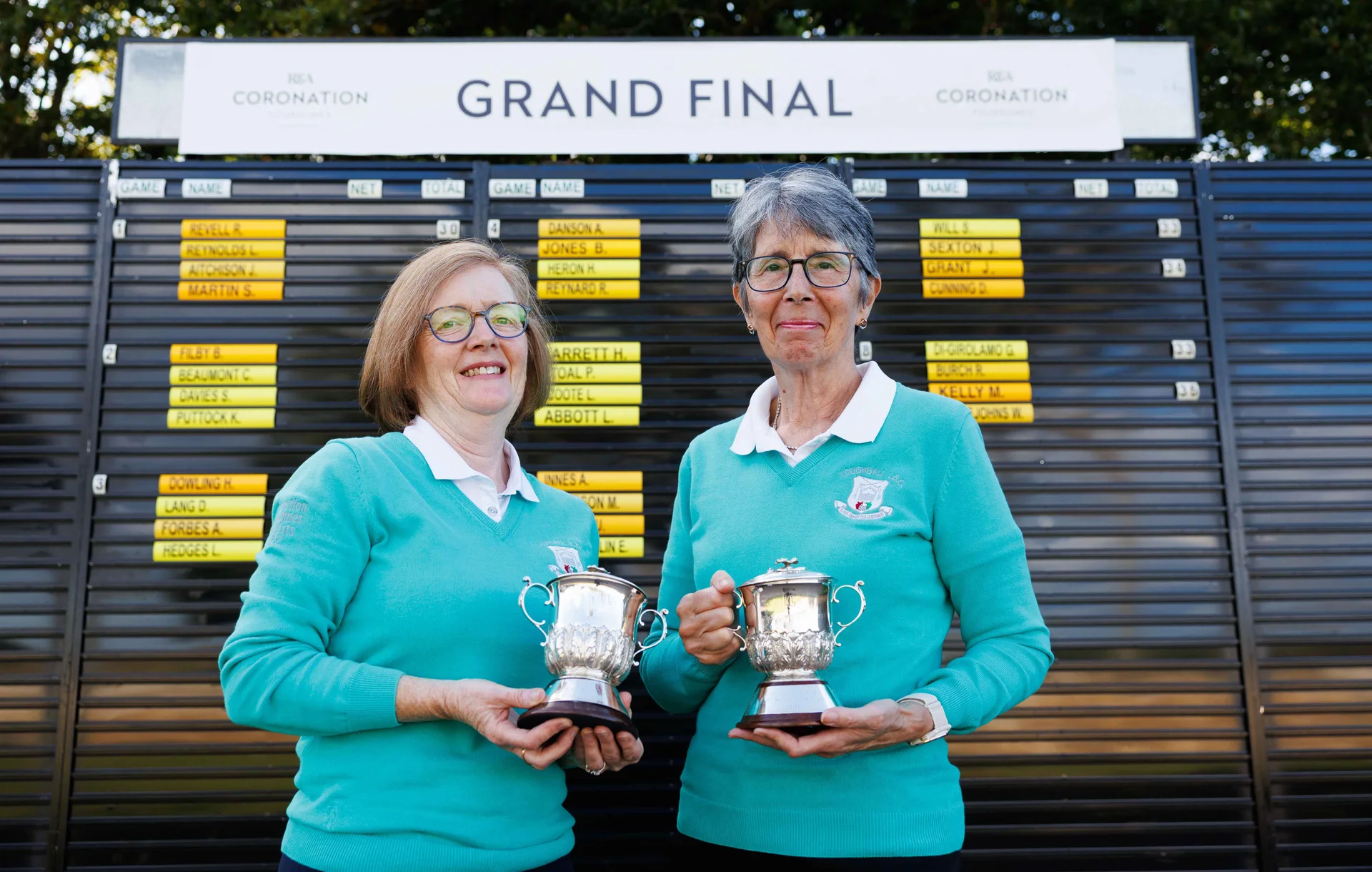 Lynn Coote and Lorraine Abbott stand in front of the scoreboard with their trophies.