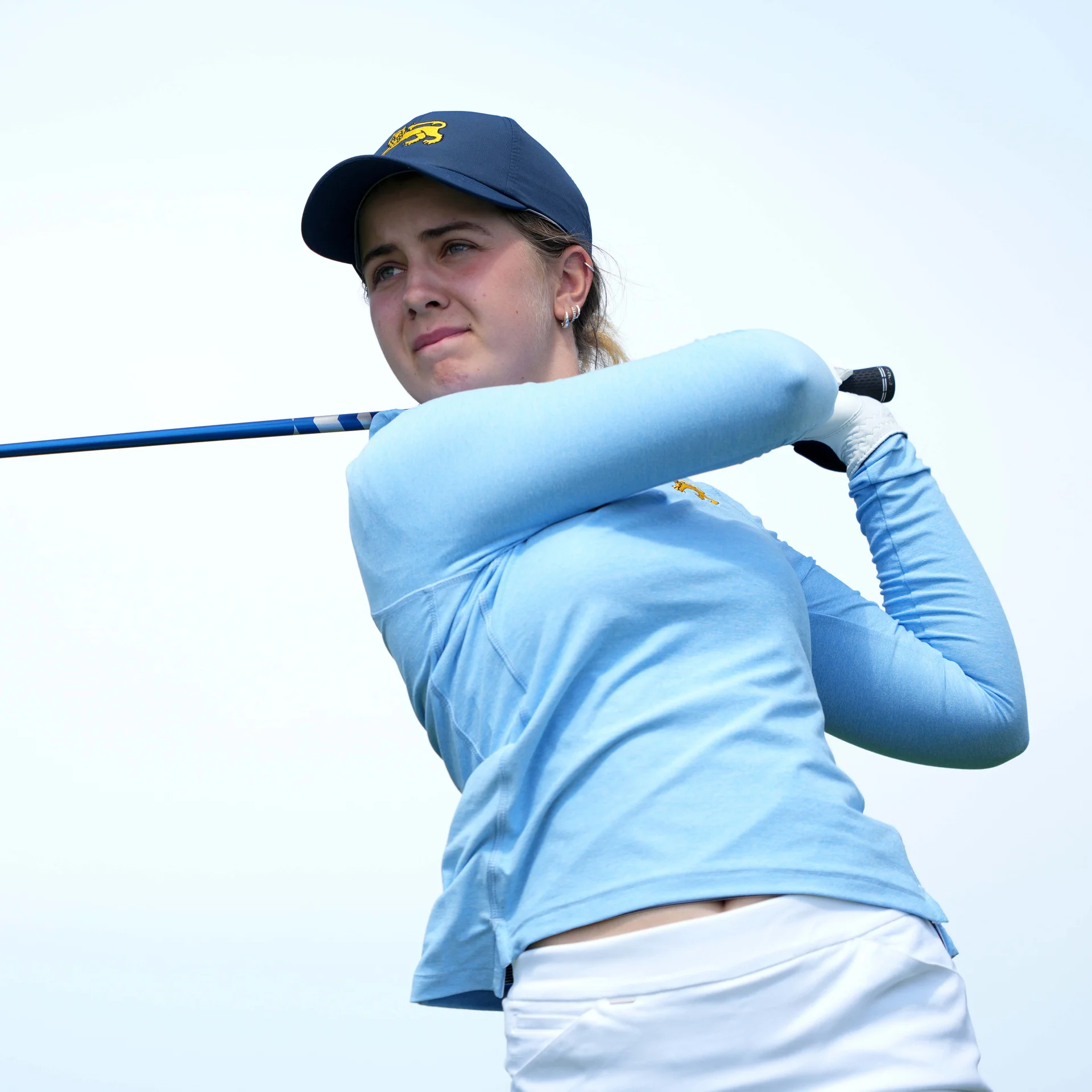 Golfer Sophia Fullbrook looks off-camera with a composed expression following a swing, wearing a light blue long-sleeve top and a navy cap with a yellow lion logo.